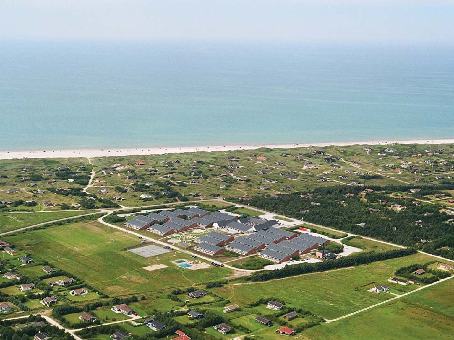 Luftfoto av et stort resortkompleks nær en sandstrand og havet, omgitt av spredte hus og grønne jorder. Landal Grønhøj Strand