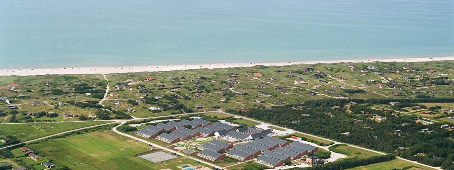 Luftfoto av et stort resortkompleks nær en sandstrand og havet, omgitt av spredte hus og grønne jorder. Landal Grønhøj Strand