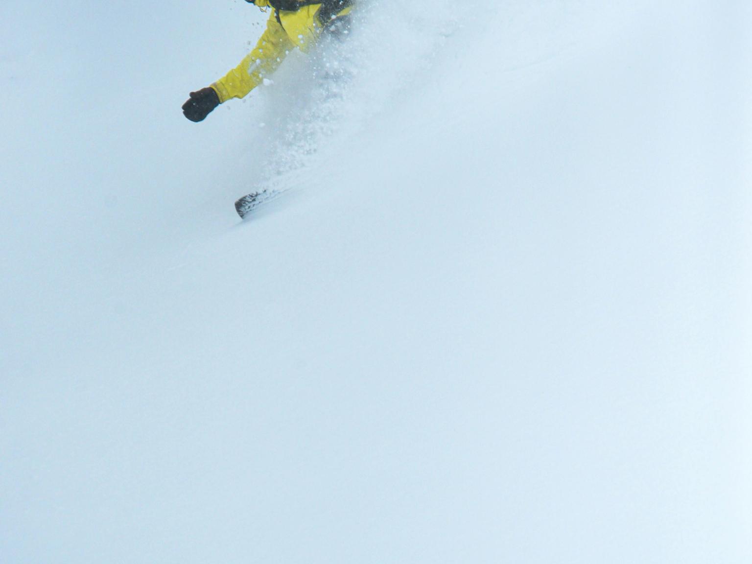 En snowboardkjører i gul jakke og blå skibriller skjærer gjennom en sving og virvler opp en stor sky av puddersnø.