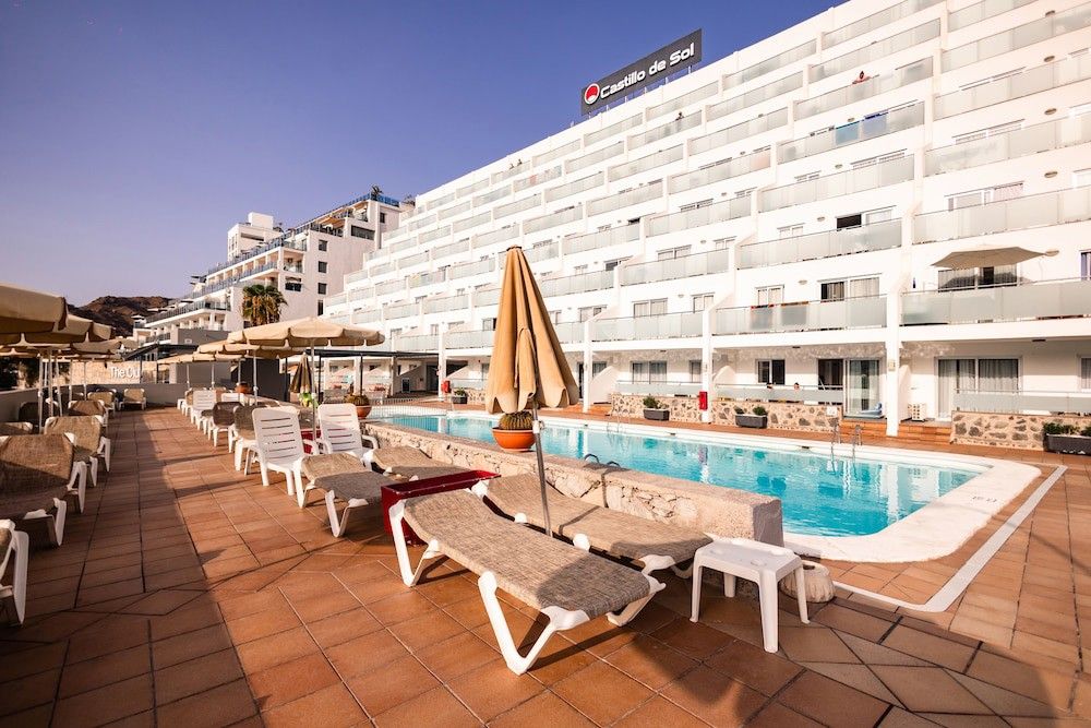 Udendørs swimmingpool med liggestole og parasoller foran den hvide Castillo de Sol hotelbygning.