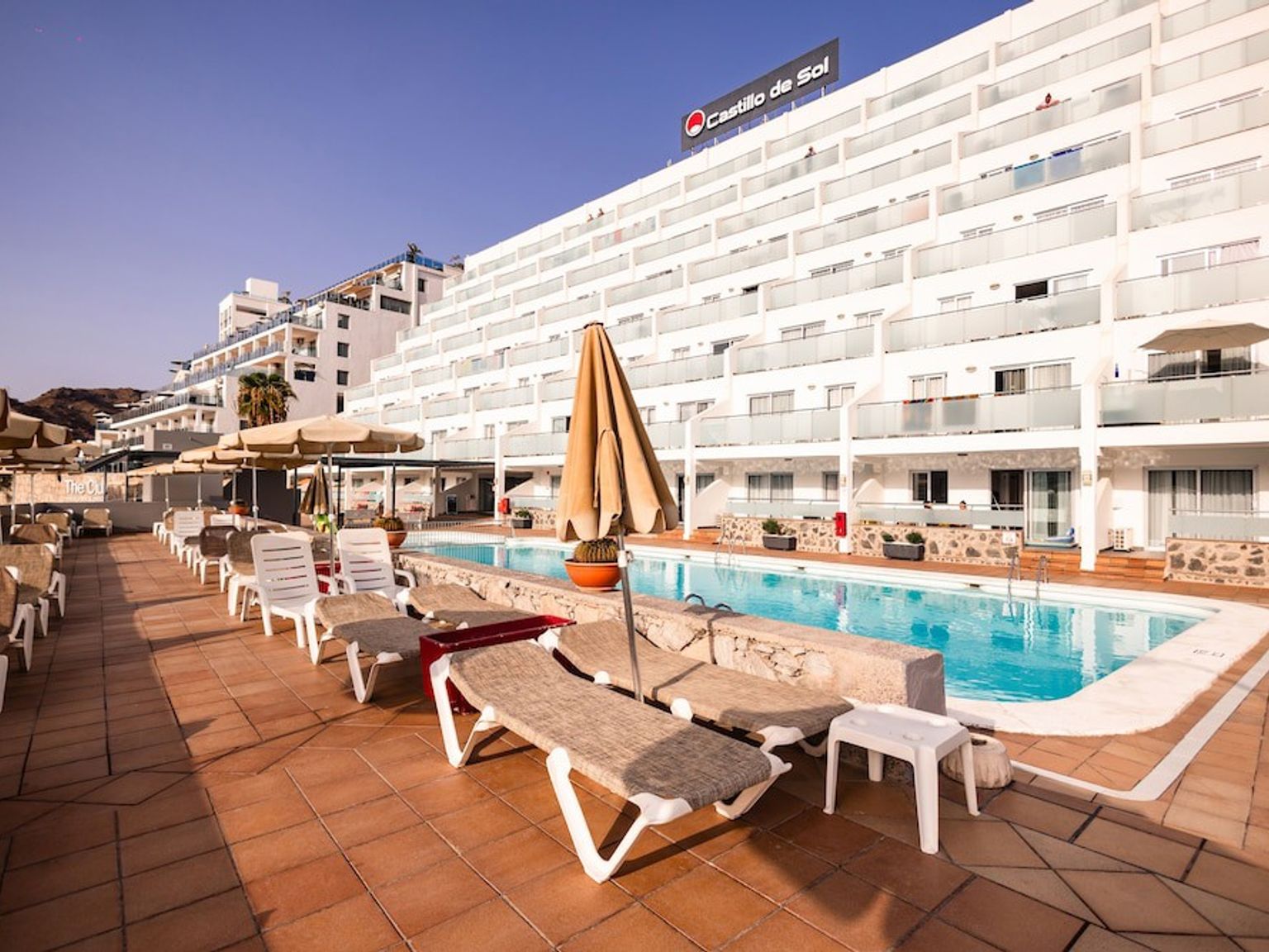 Utomhuspool med solstolar och parasoller framför det vita Castillo de Sol-hotellbyggnaden.
