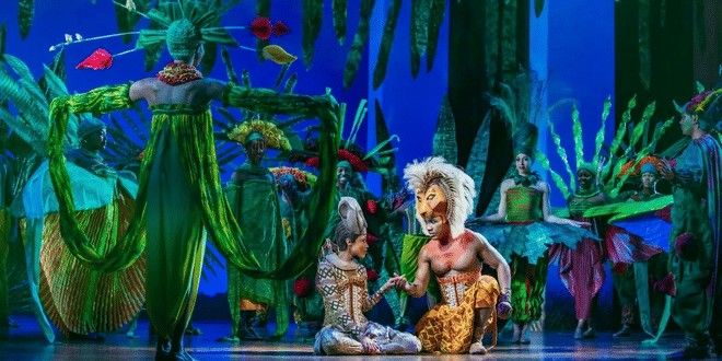 En scenuppsättning av Lejonkungen med Simba och Nala som håller varandra i handen, omgivna av artister i livfulla gröna kostymer.