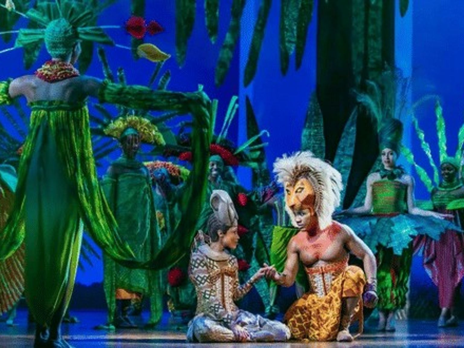 En scenuppsättning av Lejonkungen med Simba och Nala som håller varandra i handen, omgivna av artister i livfulla gröna kostymer.
