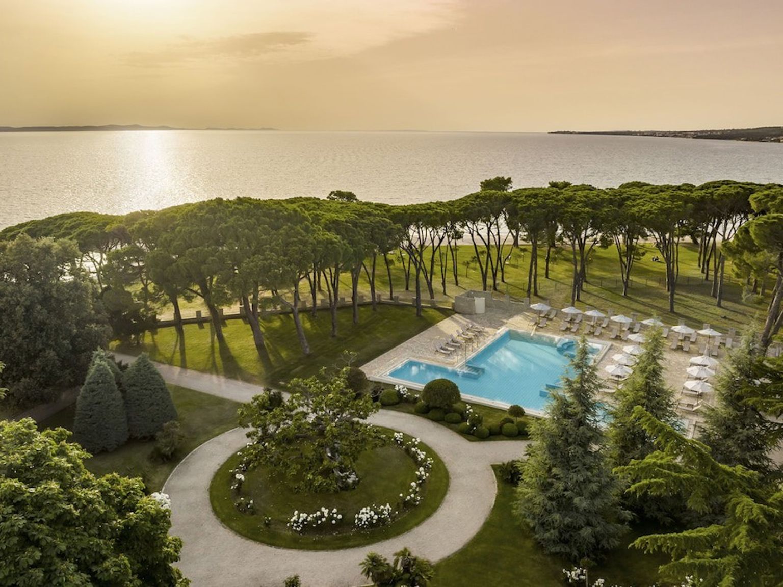 Aerial view of a large outdoor swimming pool surrounded by manicured gardens and tall trees, with the calm sea under a warm sky in the background. Falkensteiner Hotel Adriana - Adults Only