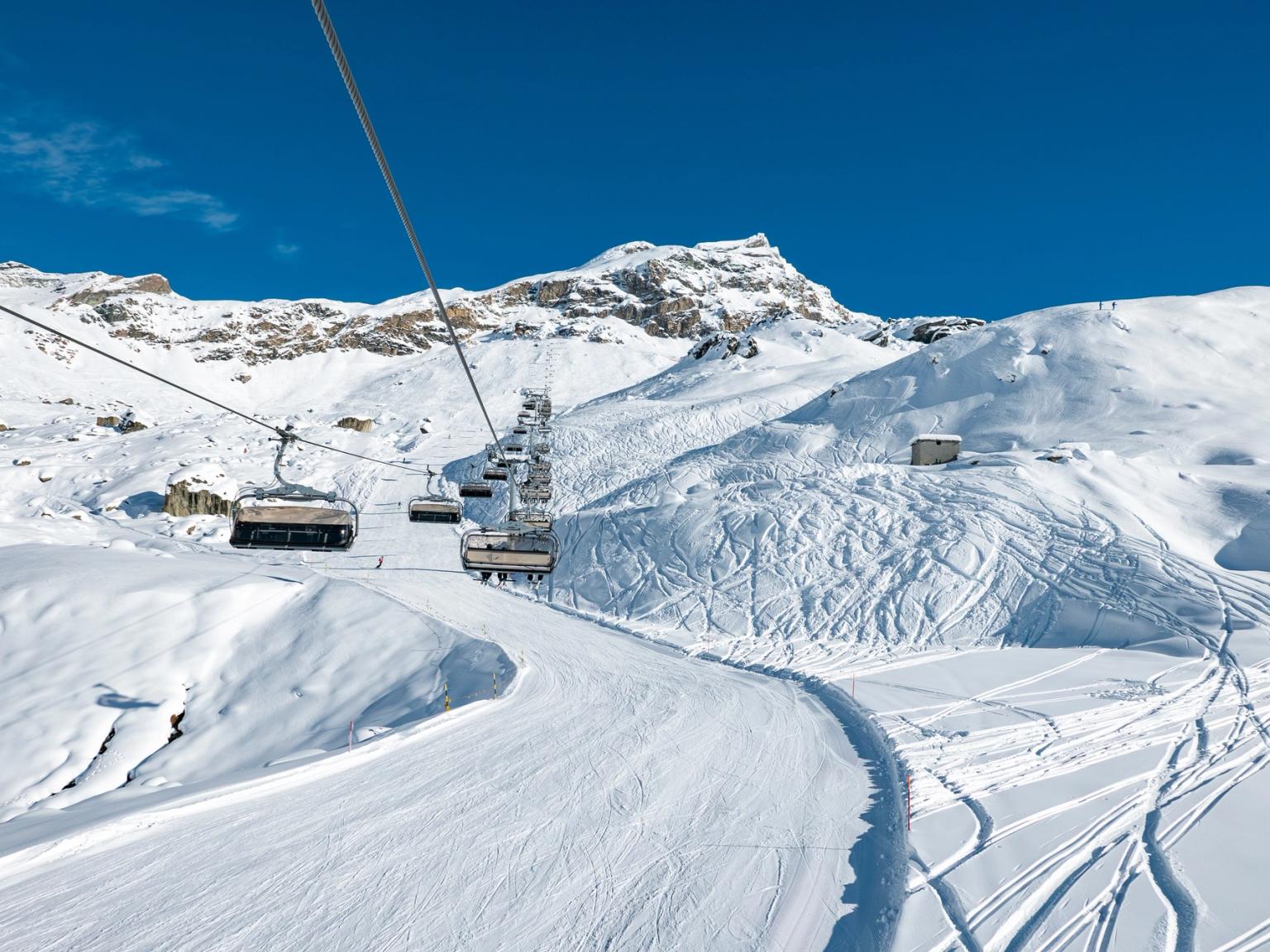 Tomme skiheisstoler beveger seg oppover en snødekt fjellside med preparerte løyper og mange skispor under en klarblå himmel.