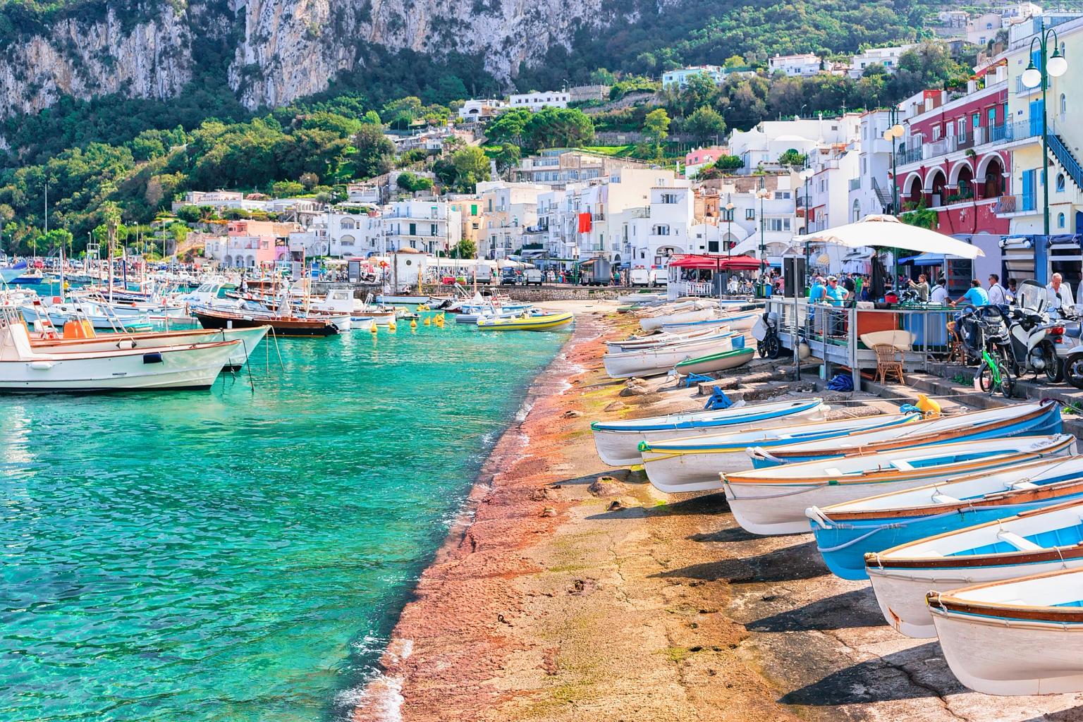 Een levendige havenscène op Capri met turkoois water, boten die zijn afgemeerd en op het strand zijn getrokken, en een kleurrijke stad tegen de kliffen.