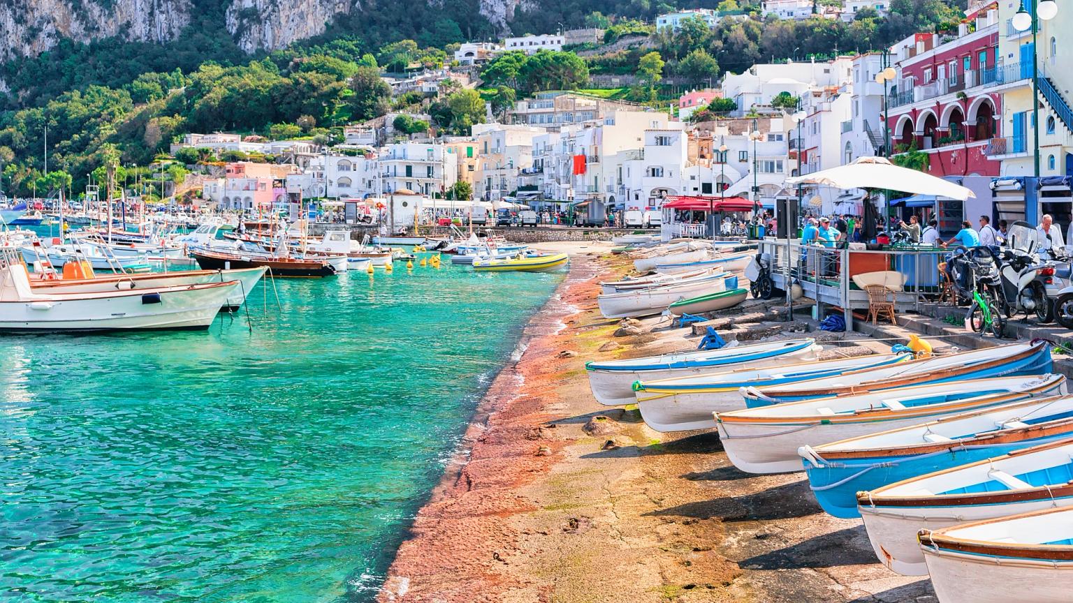 Een levendige havenscène op Capri met turkoois water, boten die zijn afgemeerd en op het strand zijn getrokken, en een kleurrijke stad tegen de kliffen.