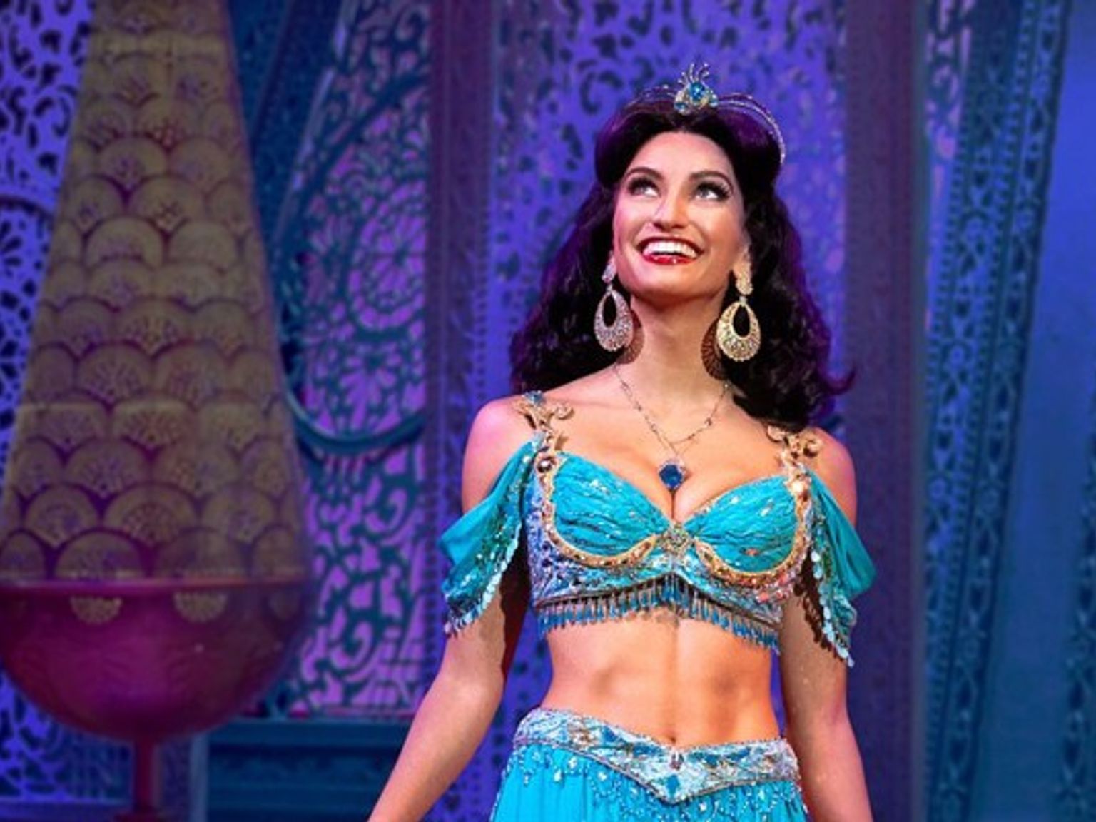 Une femme portant un costume turquoise de la princesse Jasmine sourit et regarde vers la scène.