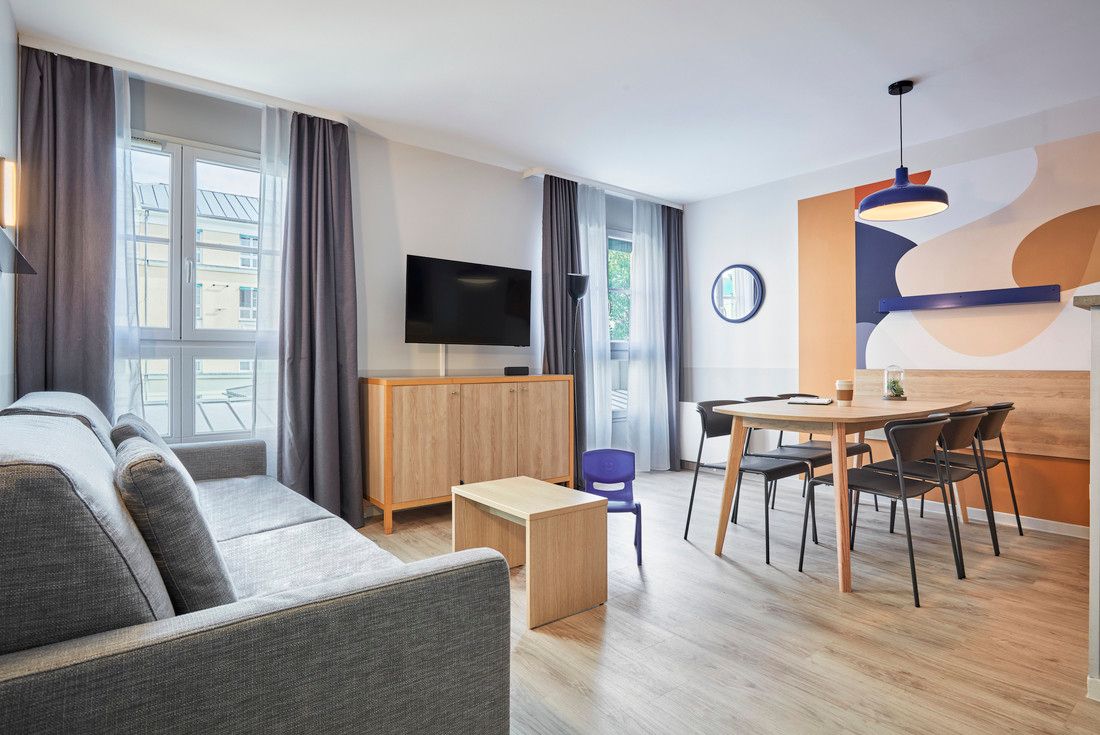 En lys, moderne åpen planløsning stue og spisestue med en grå sofa, lette tremøbler, lyse gulv og en aksentvegg med abstrakte oransje og blå former.