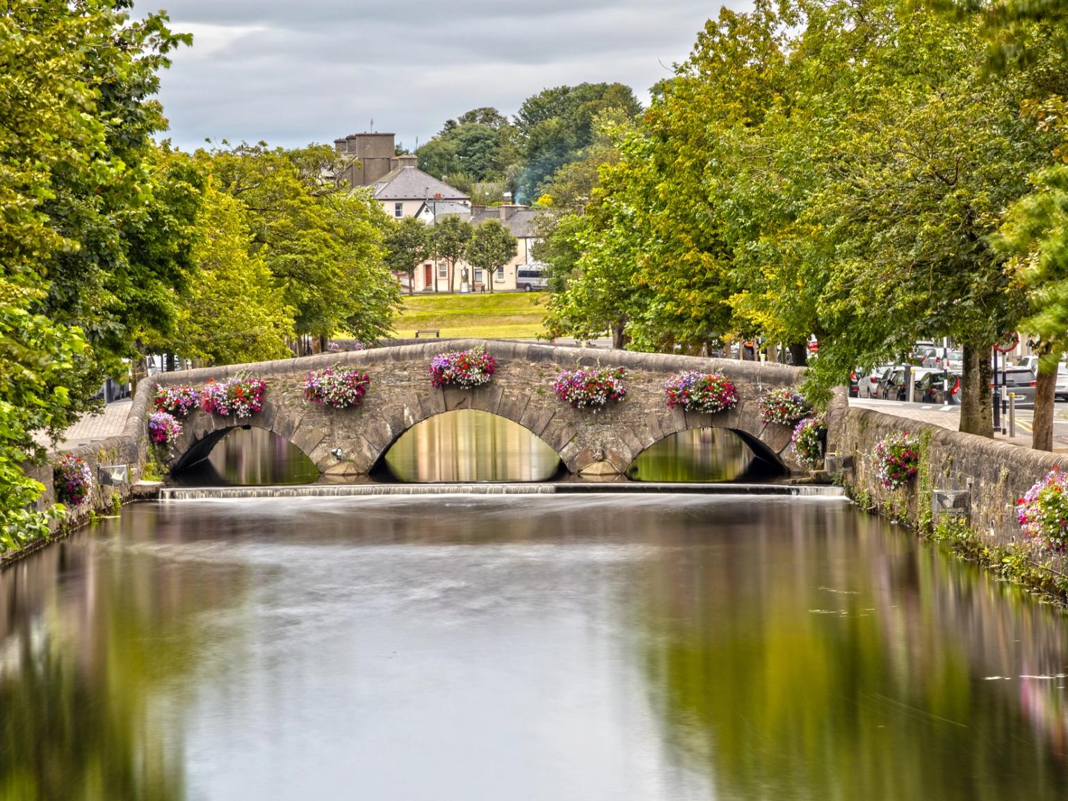 Stone arch bridge with colorful flower baskets spanning a calm river, flanked by green trees.