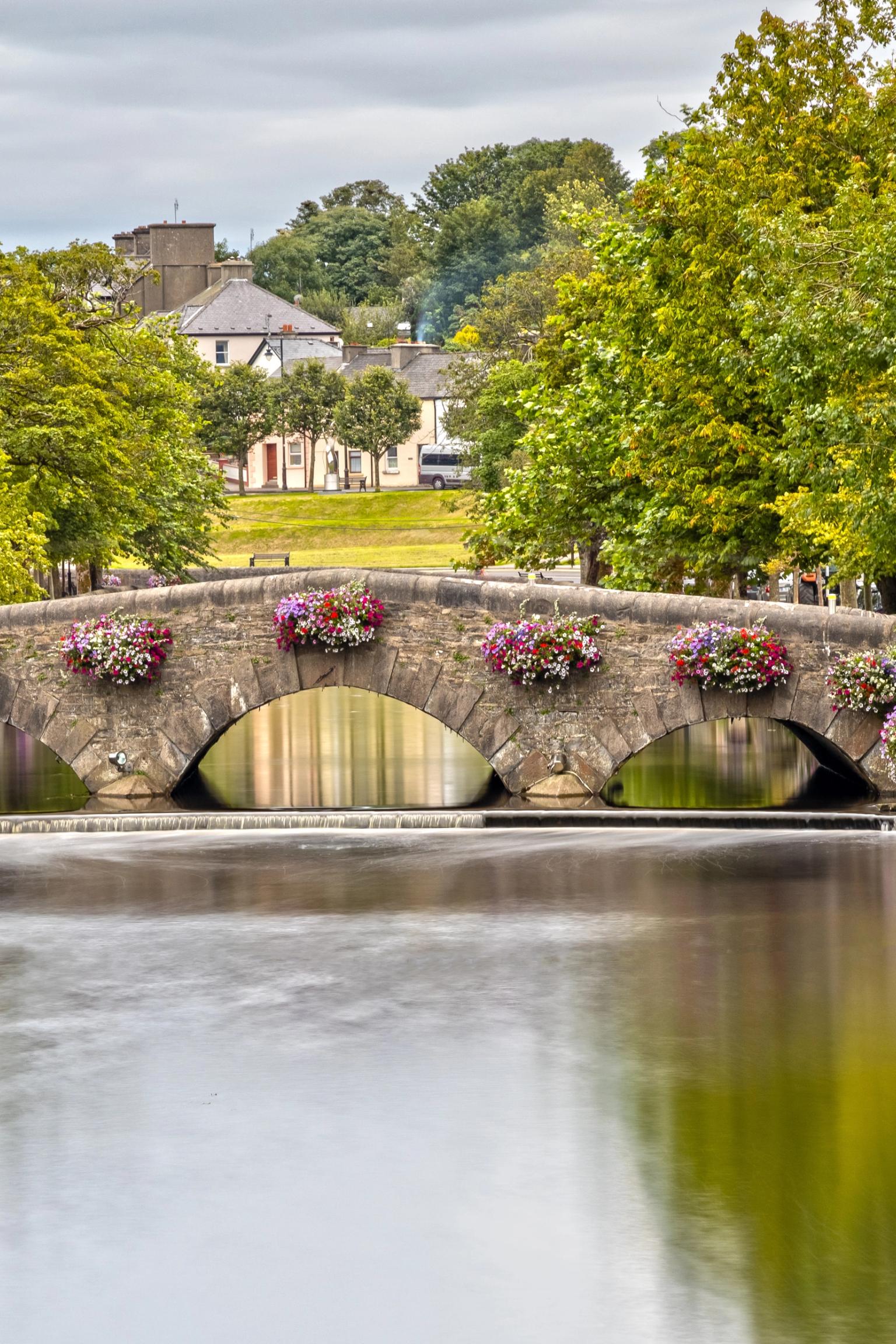 Stone arch bridge with colorful flower baskets spanning a calm river, flanked by green trees.