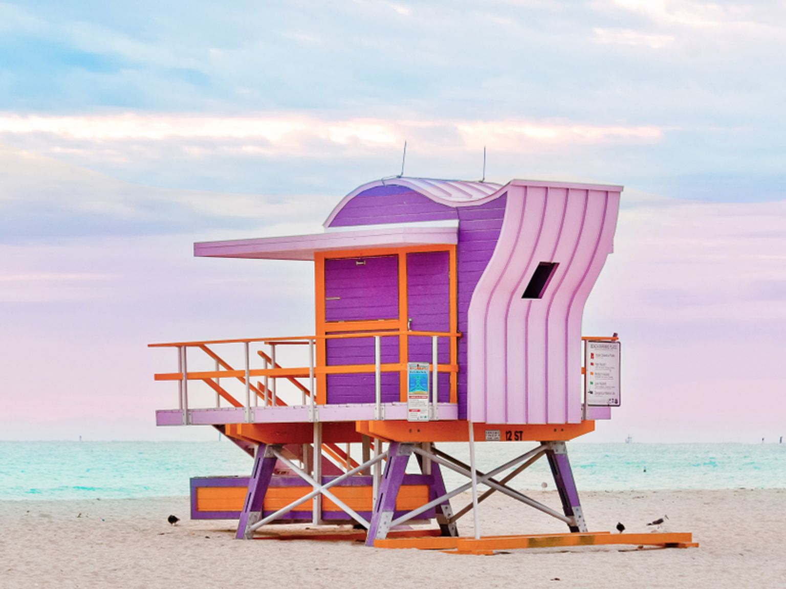 Et lilla, orange og pink livreddertårn på en sandstrand under en pastelhimmel. Miami, USA