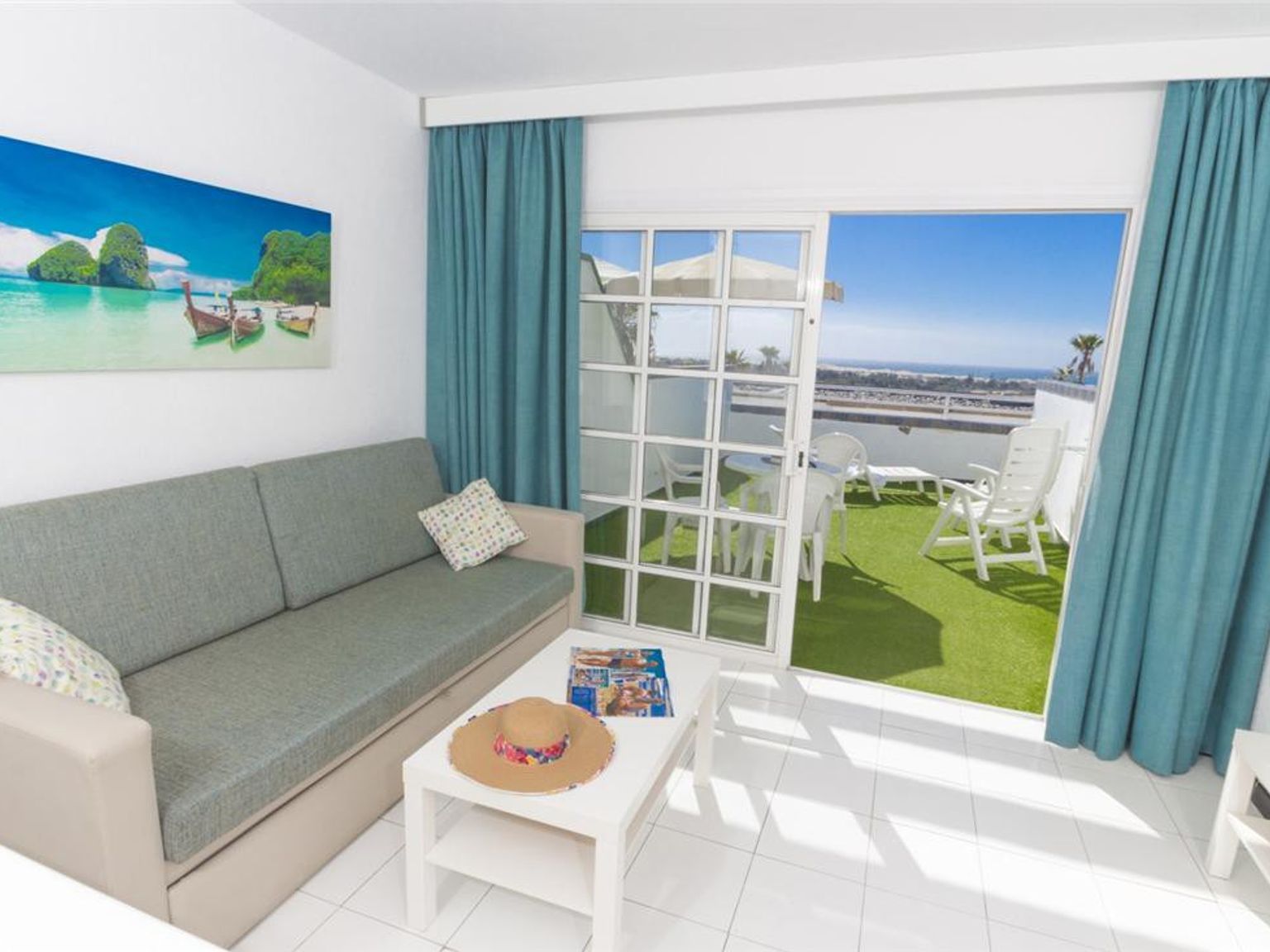 En lys stue med en sofa, strandkunst og en åpen dør til en balkong med kunstgress og havutsikt.