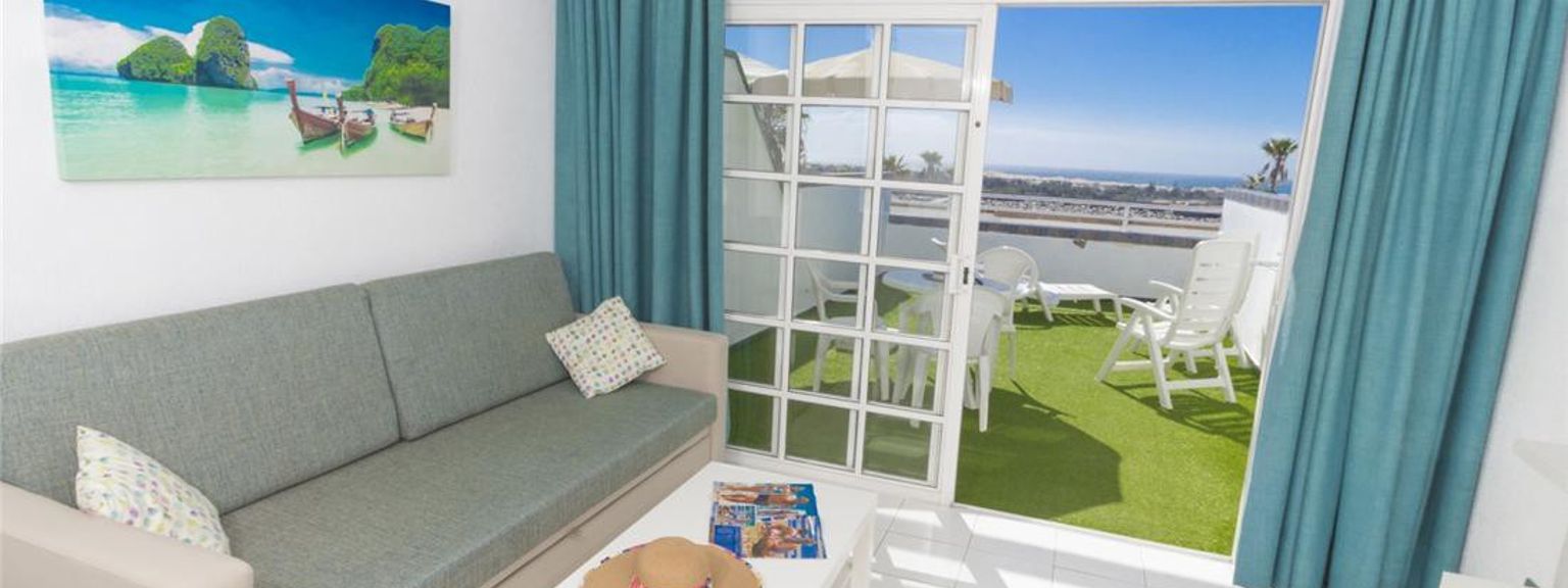 En lys stue med en sofa, strandkunst og en åpen dør til en balkong med kunstgress og havutsikt.