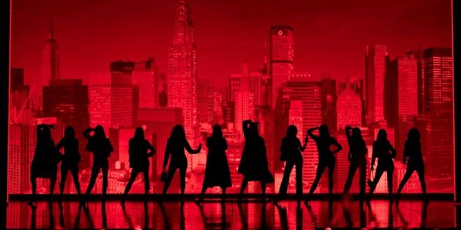 Eleven silhouetted figures pose on a reflective floor in front of a red city skyline.