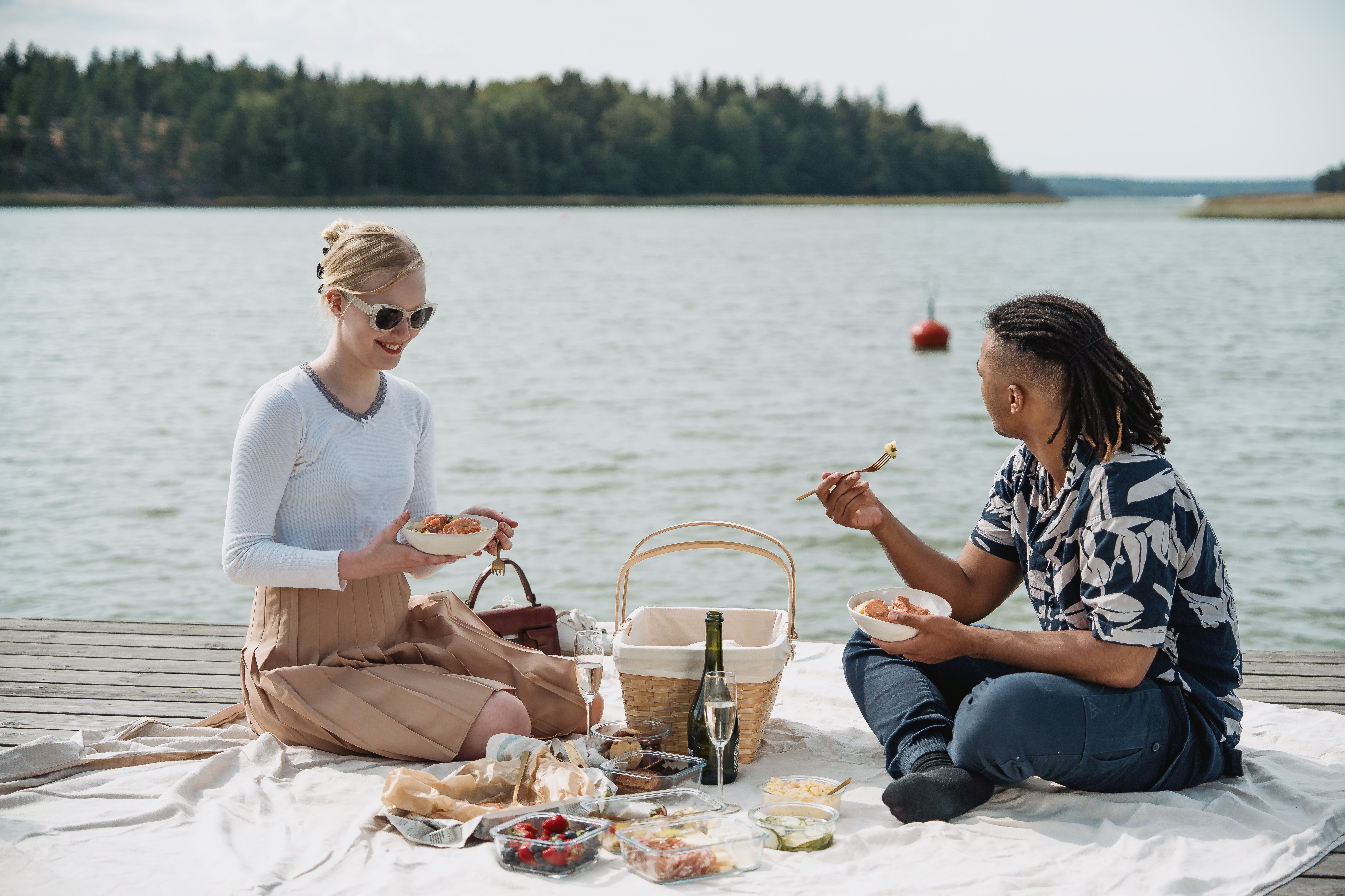 En person står på en klippefyldt kyst og vender sig mod den klare sol over et glitrende hav under en blå himmel.