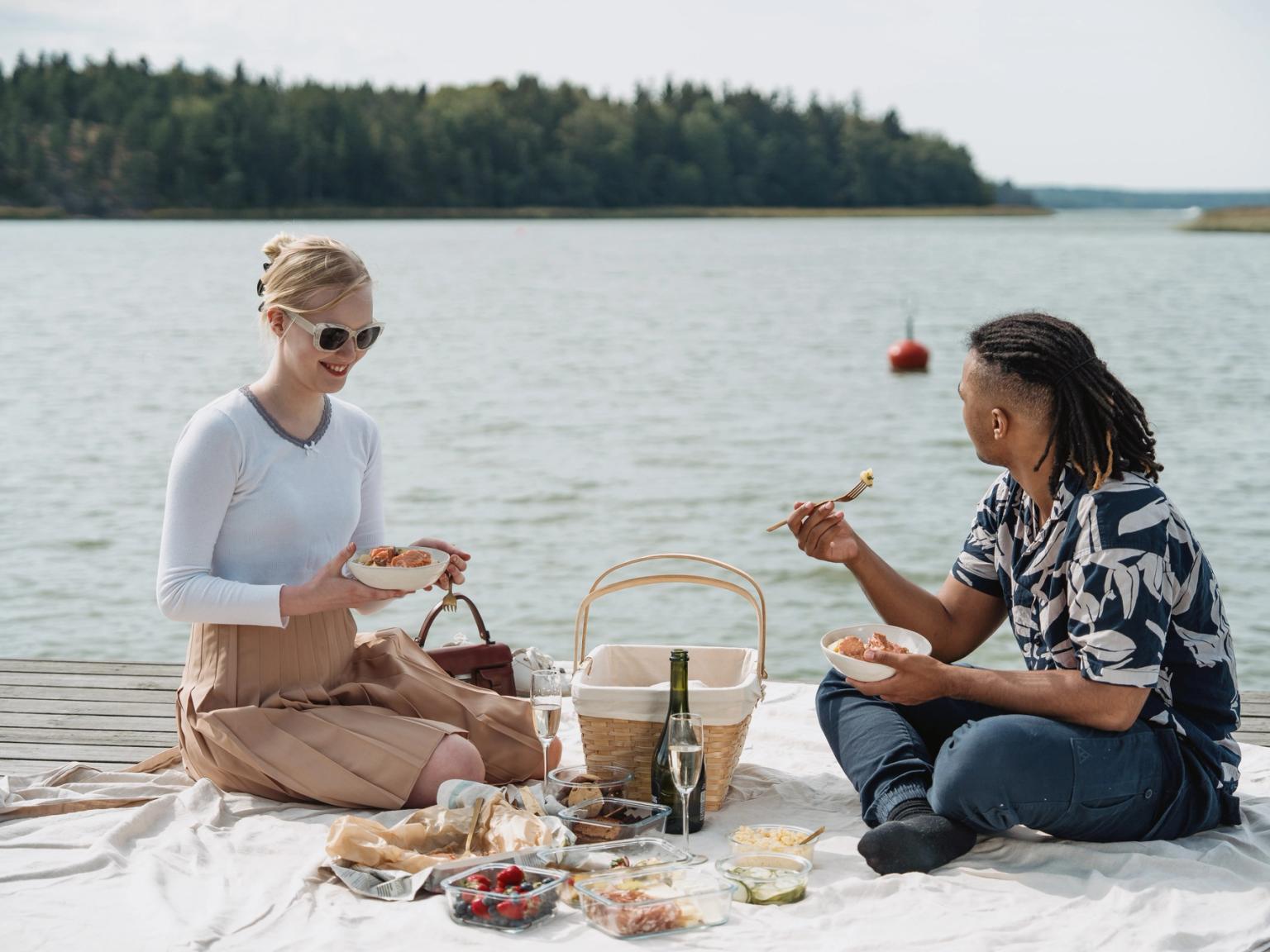 En person står på en klippefyldt kyst og vender sig mod den klare sol over et glitrende hav under en blå himmel.