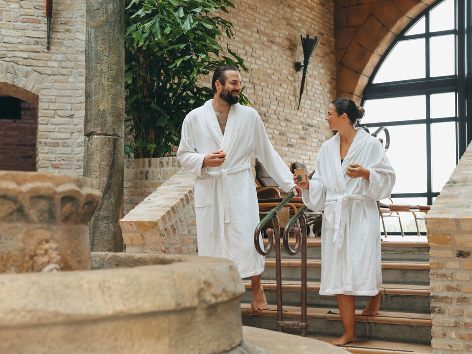 Par i hvite badekåper som går ned steintrapper på et spa, smilende til hverandre.