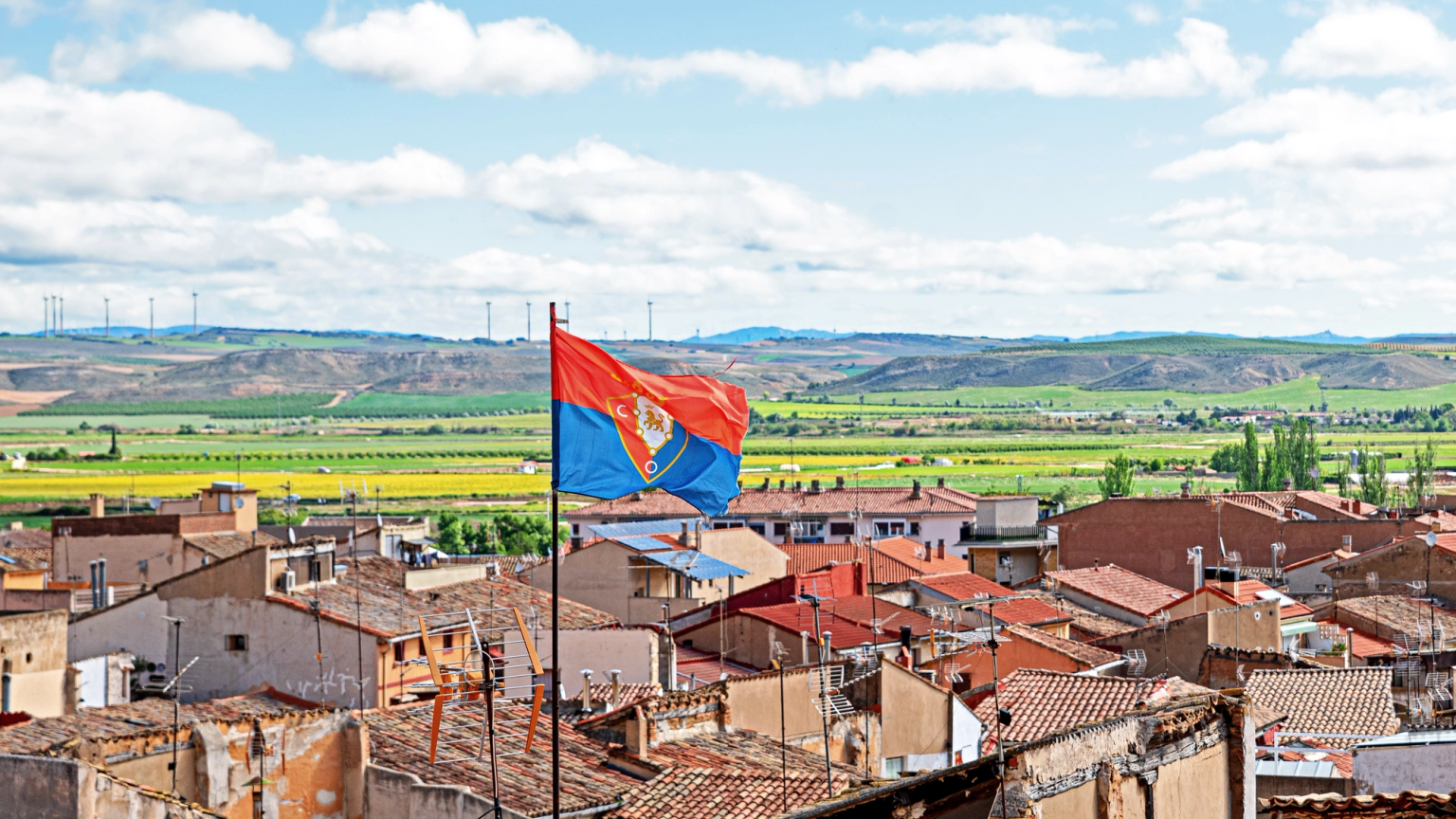 A red and blue flag with a crest flies over a town with terracotta roofs, against a backdrop of green fields, distant hills with wind turbines, and a cloudy sky.