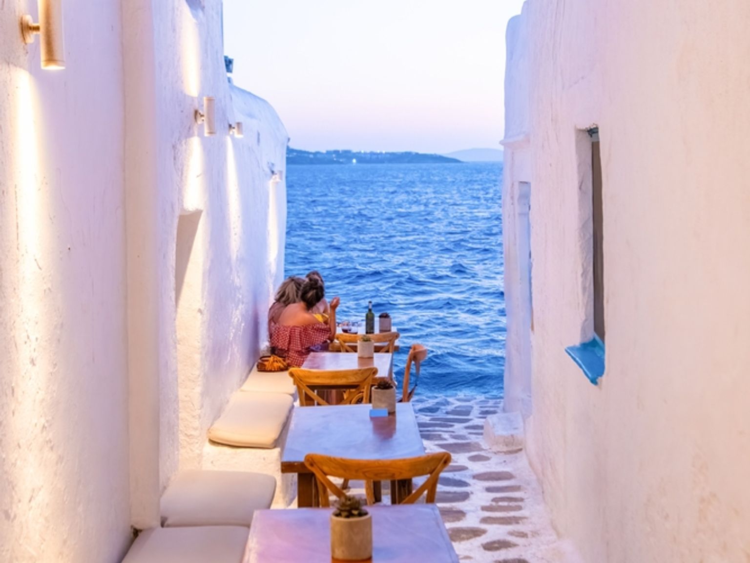 Smal hvit smug med uteserveringsbord som leder mot havutsikt. Mykonos, Hellas