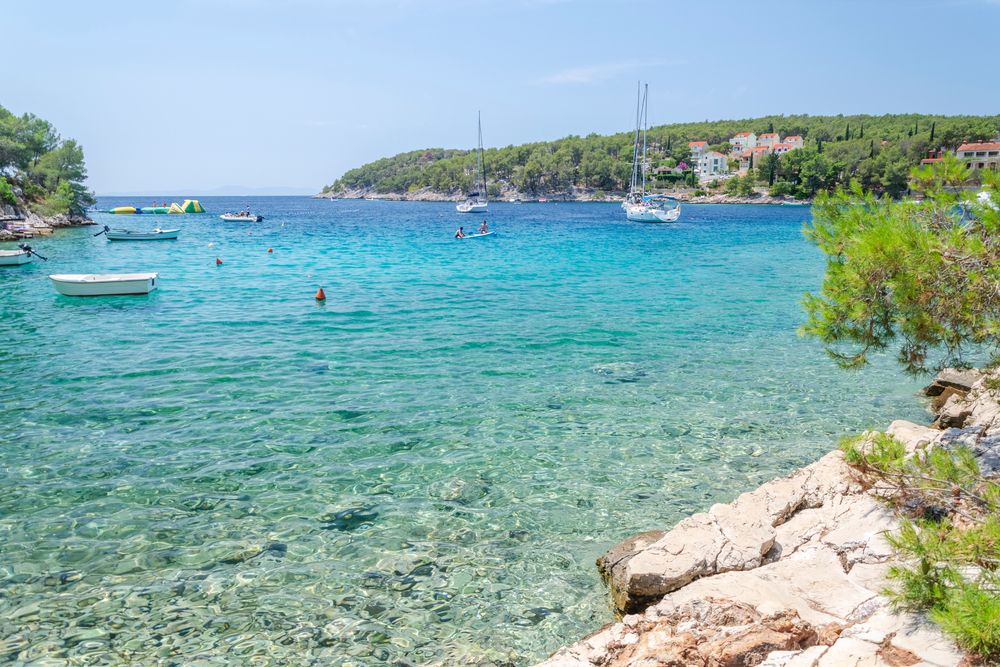 Klar turkis bugt med både, skovklædte bakker, huse og en klippefyldt forgrund. Milna, Brac