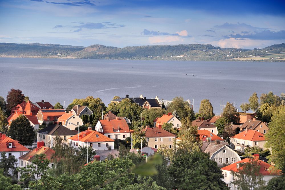 En landsby med rød takede hus ved en stor innsjø og fjerne åser under en blå himmel.