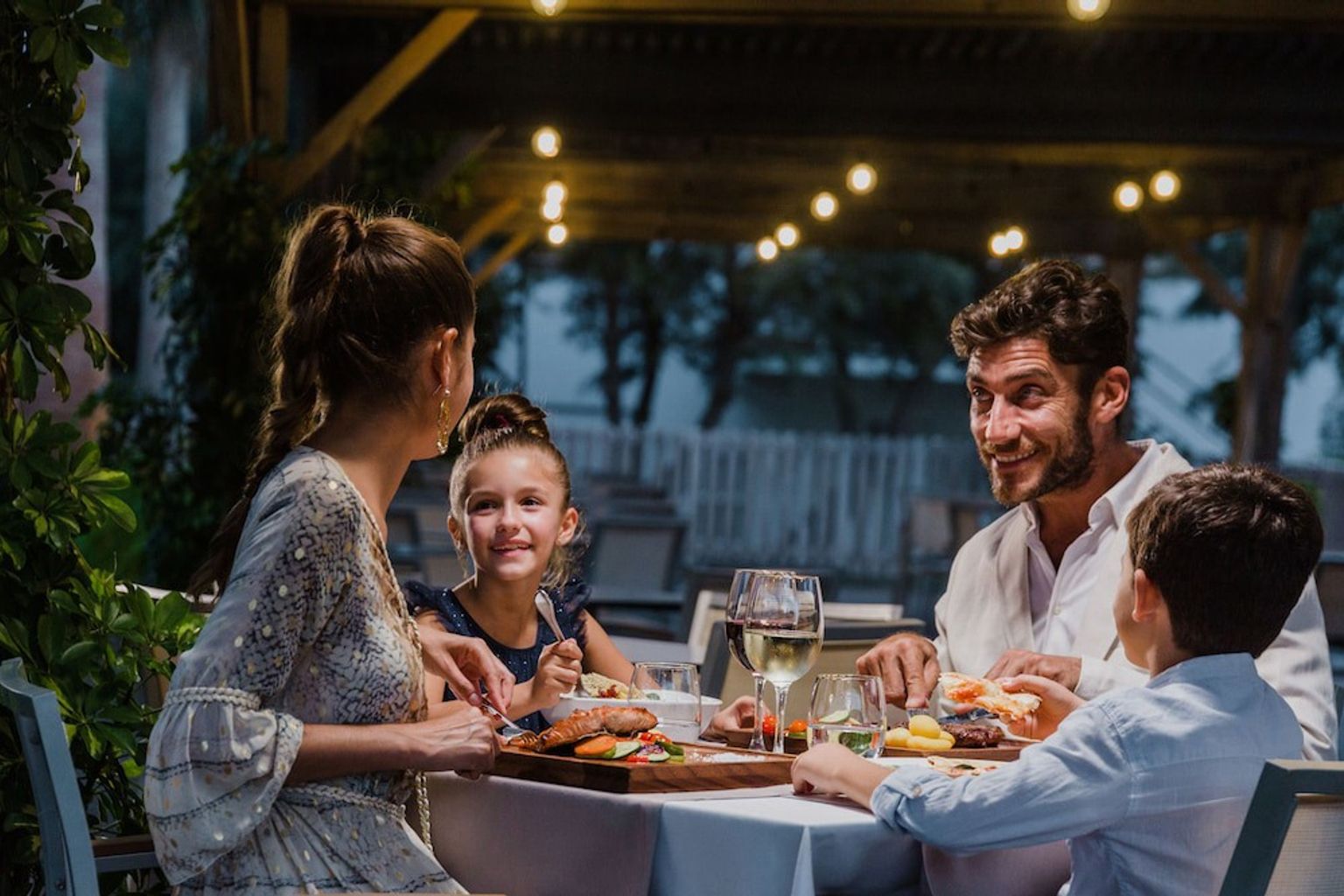 En familj på fyra njuter av en middag utomhus på kvällen under ljusslingor. Aparthotel Club del Sol Resort & SPA