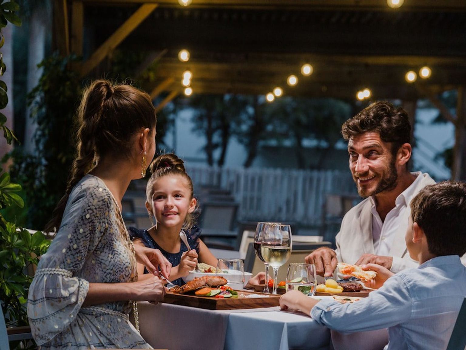Eine vierköpfige Familie genießt abends ein Abendessen im Freien unter Lichterketten. Aparthotel Club del Sol Resort & SPA