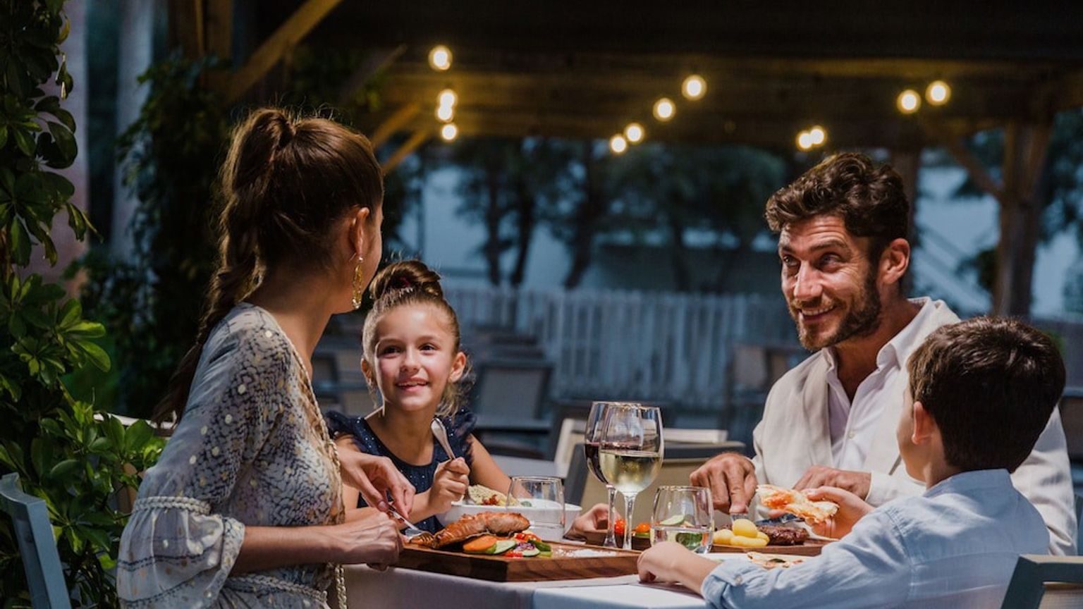 En familj på fyra njuter av en middag utomhus på kvällen under ljusslingor. Aparthotel Club del Sol Resort & SPA