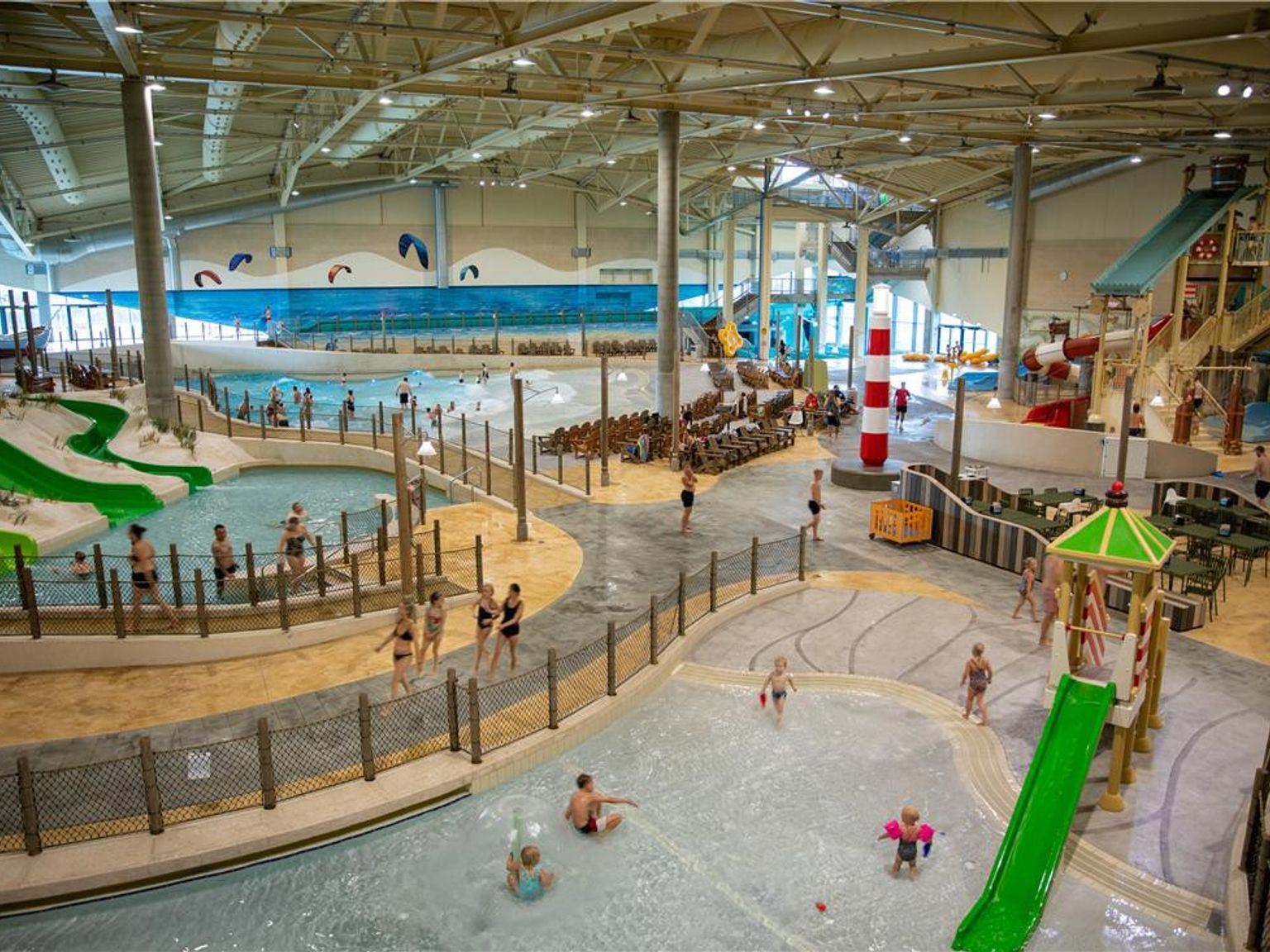 En luftfoto af en travl indendørs vandland med forskellige pools, vandrutsjebaner, en bølgebassin, et rød og hvidt fyrtårn og mange mennesker, der nyder attraktionerne. Lalandia Resort Søndervig