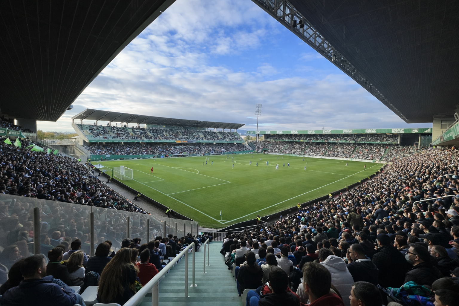 A large football stadium packed with fans watching a match on a green field under a partly cloudy sky.