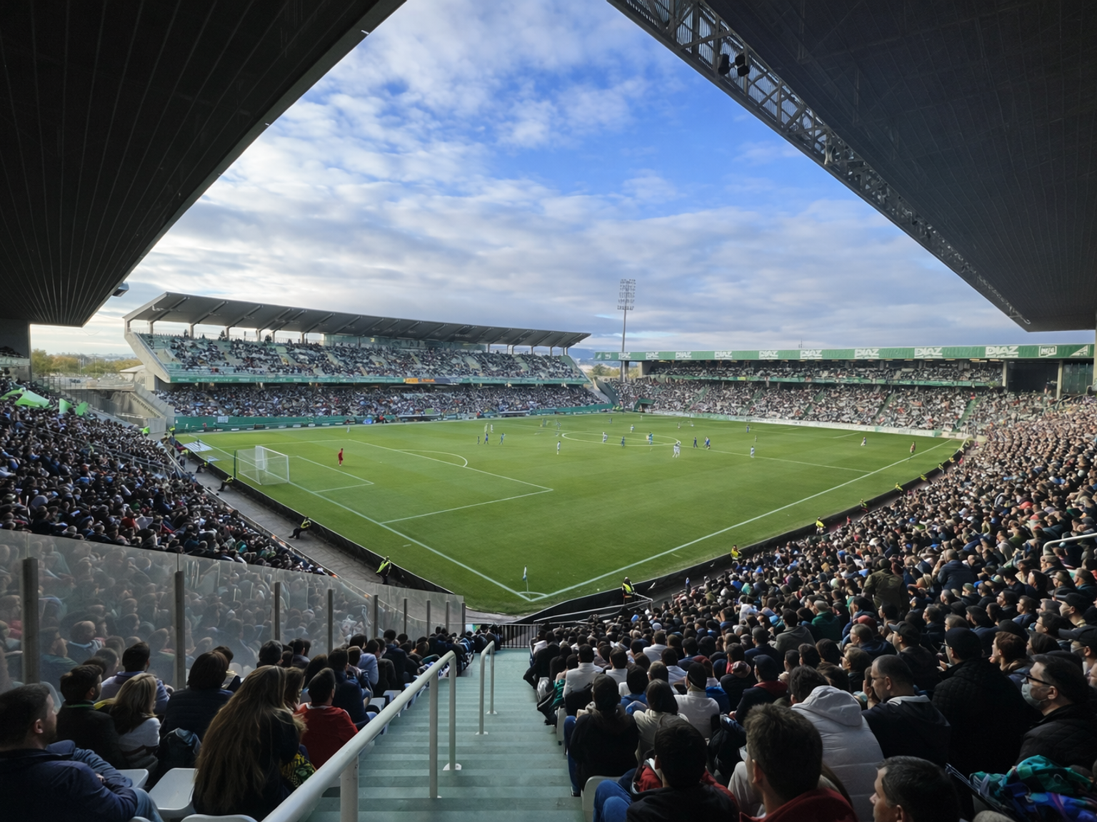 A large football stadium packed with fans watching a match on a green field under a partly cloudy sky.