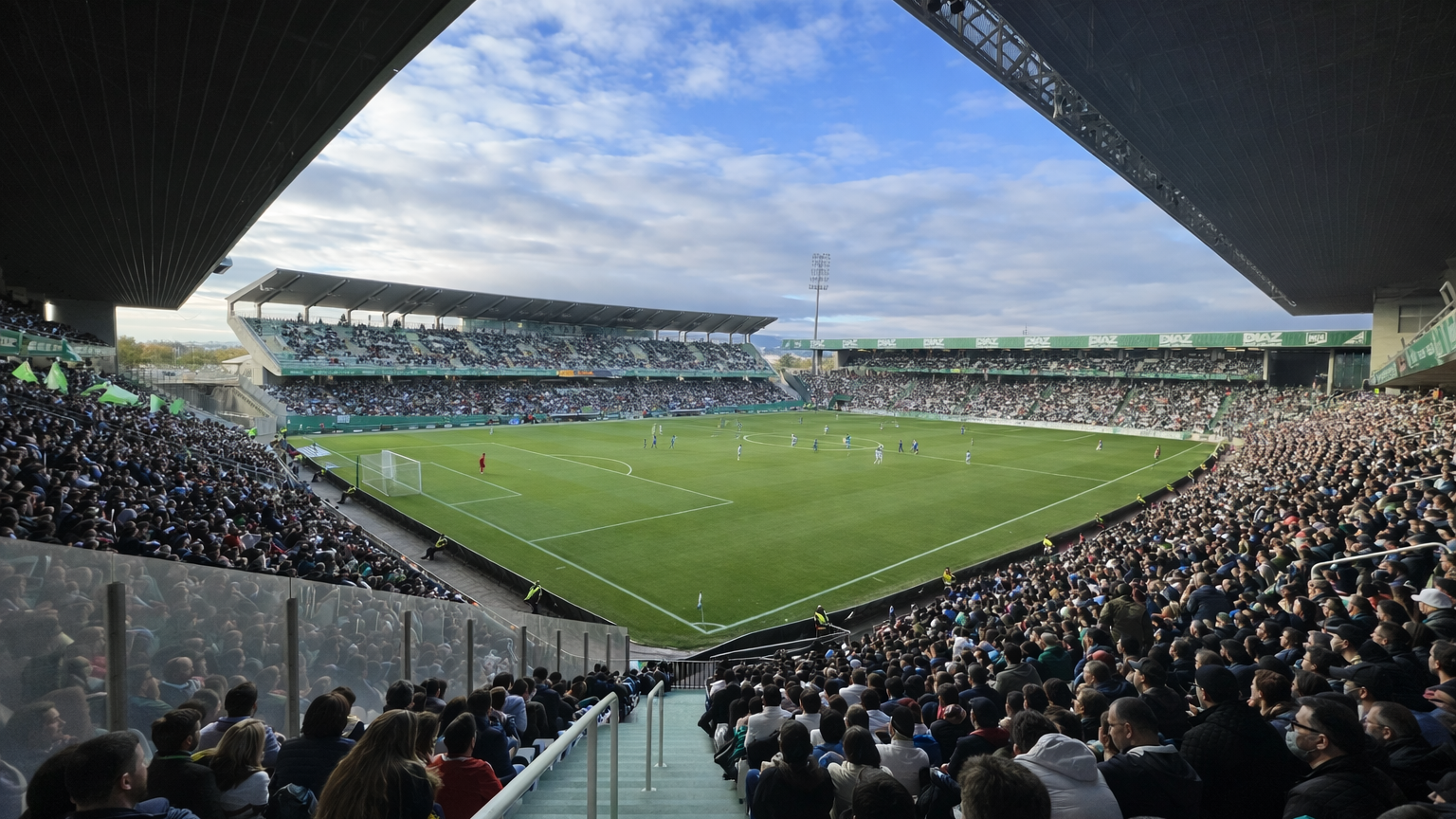 A large football stadium packed with fans watching a match on a green field under a partly cloudy sky.
