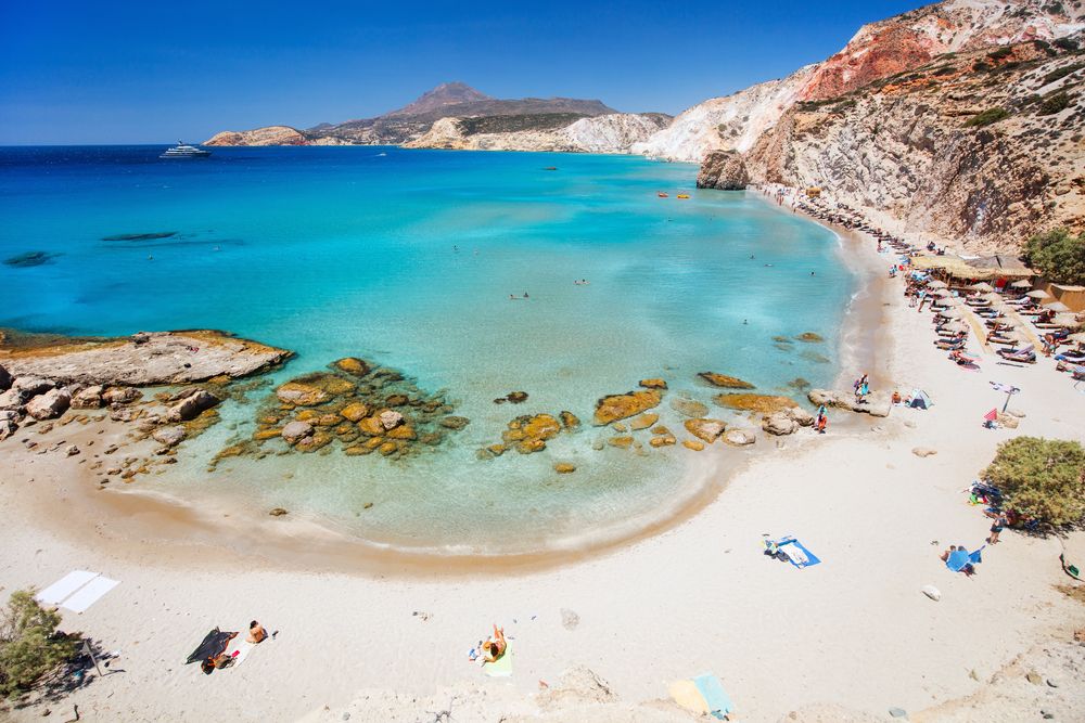 A vibrant white sand beach with clear turquoise water, colorful rock formations, and people enjoying the sun. Milos, Greece