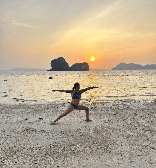 Kvinna i yoga-positionen Krigare II på en strand vid solnedgången med öar i bakgrunden.