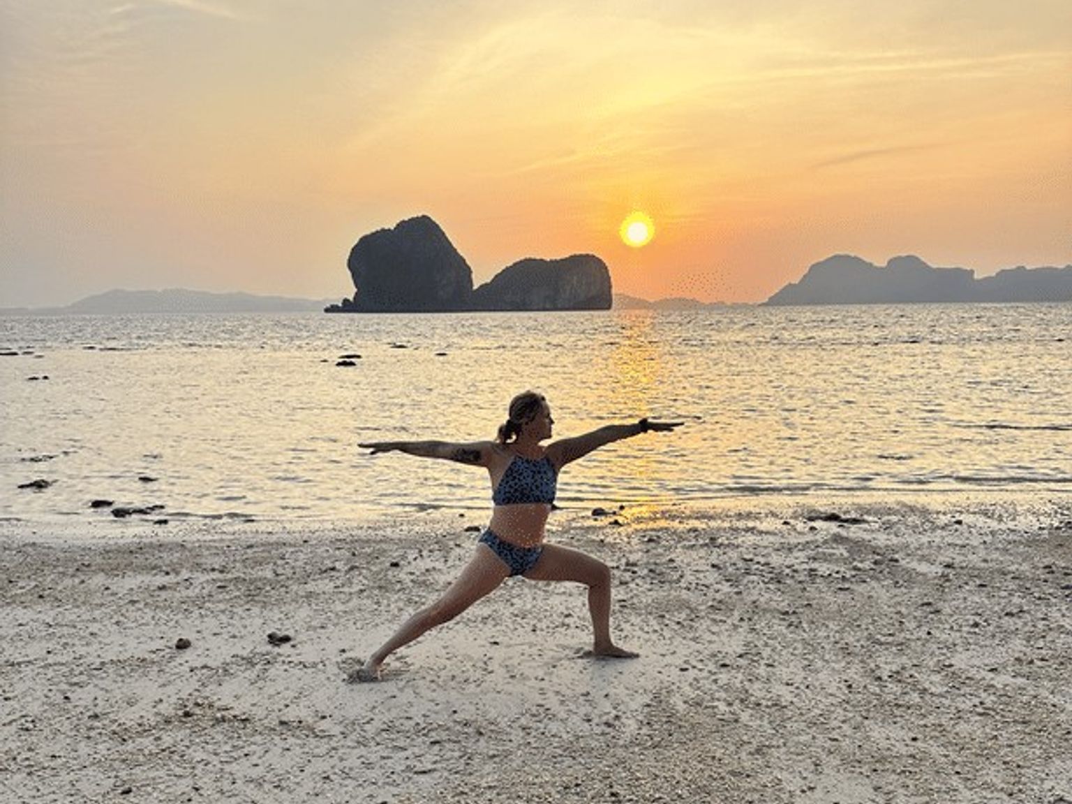 Kvinna i yoga-positionen Krigare II på en strand vid solnedgången med öar i bakgrunden.