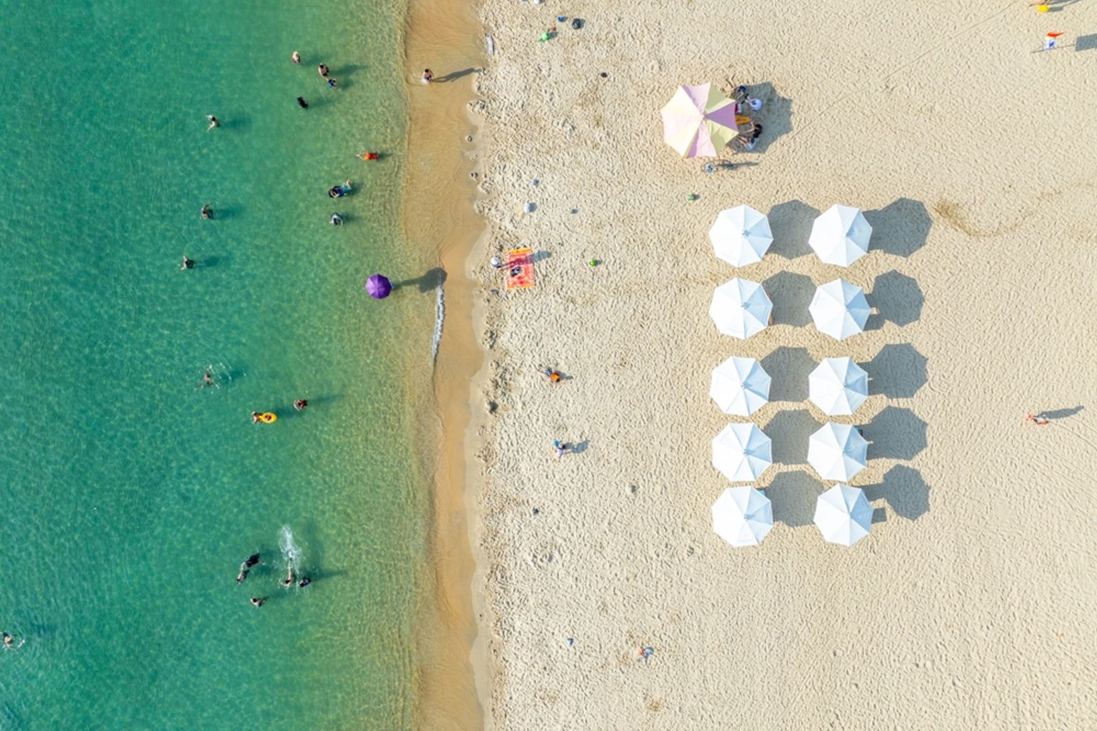 Luftfoto af en solrig strand med klart turkis vand, mange svømmere og rækker af hvide parasoller på sandet. Nha Trang, Vietnam