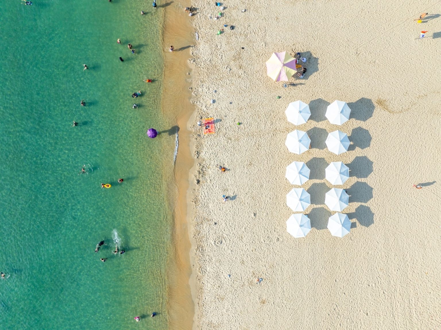 Flygvy av en solig strand med klart turkost vatten, många simmare och rader av vita parasoller på sanden. Nha Trang, Vietnam