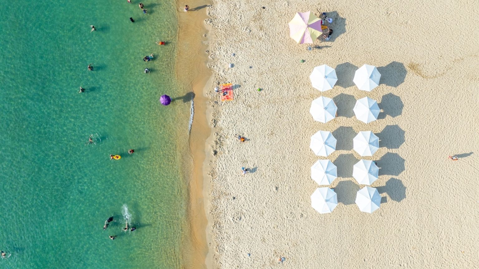 Luftfoto af en solrig strand med klart turkis vand, mange svømmere og rækker af hvide parasoller på sandet. Nha Trang, Vietnam