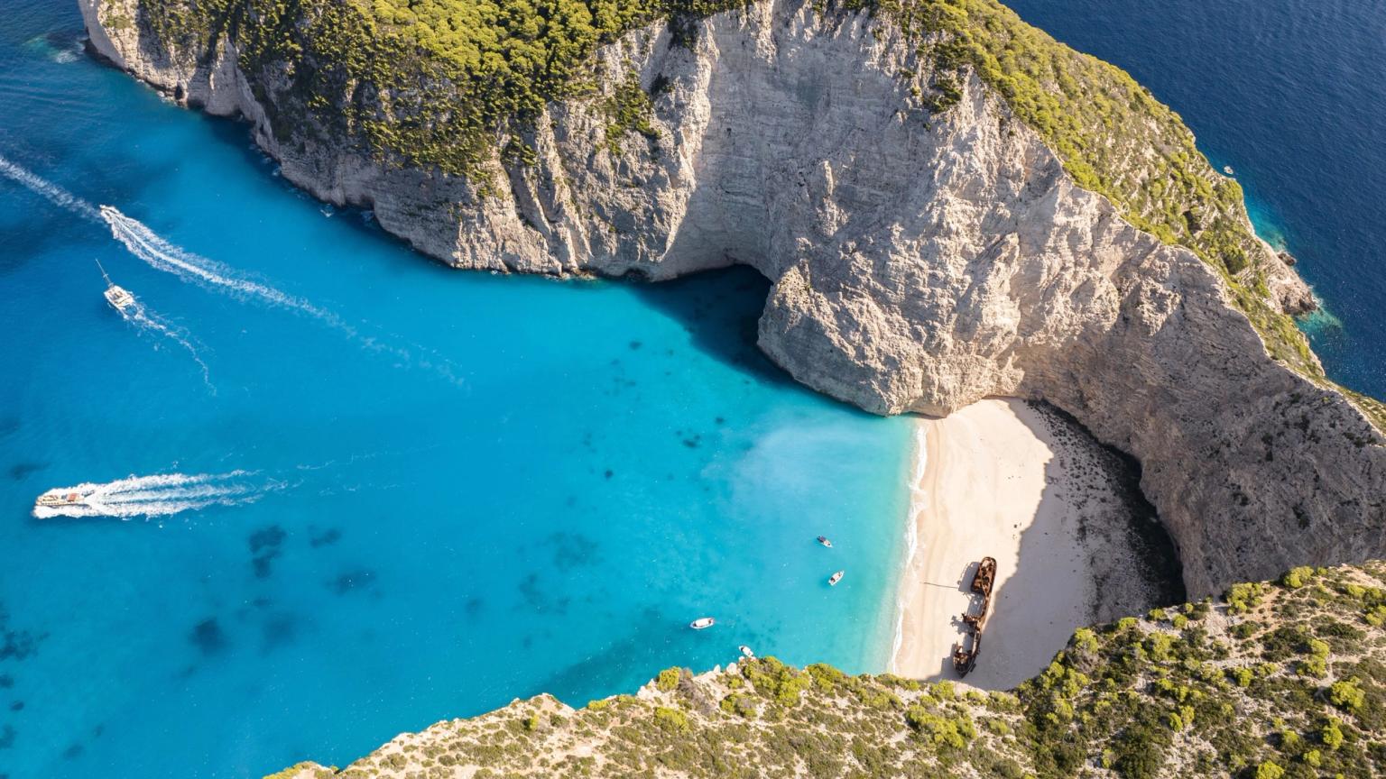 Flygfoto av en turkos bukt omgiven av vita klippor, med en sandstrand och ett skeppsvrak. Zakynthos