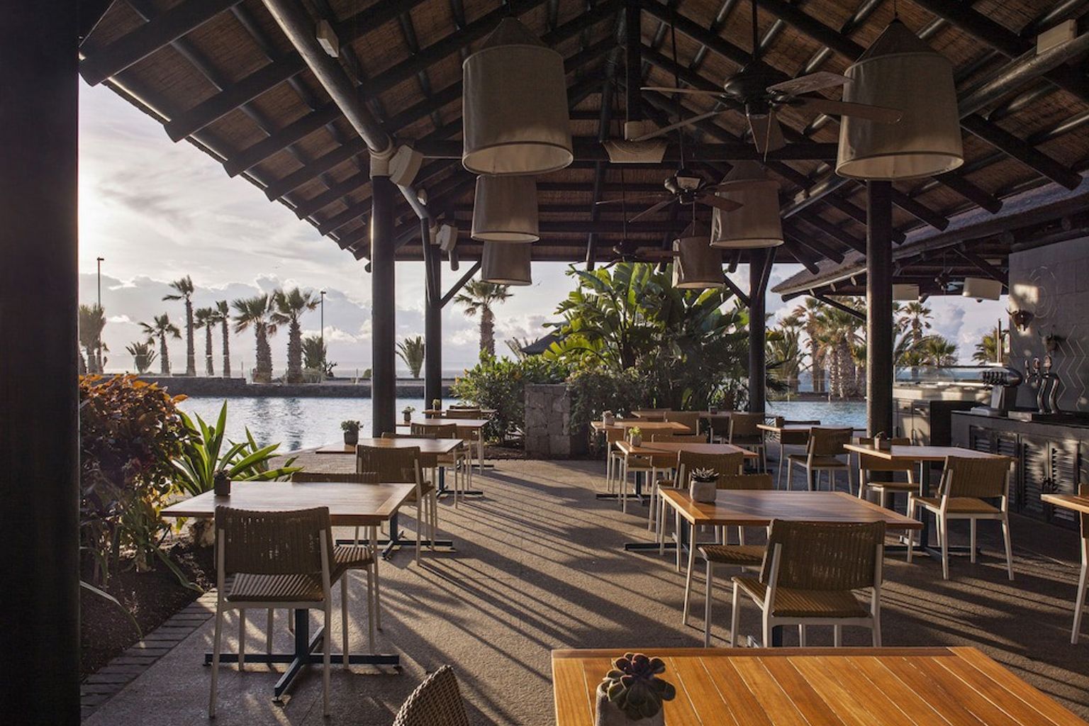 en restaurang med bord och stolar under ett halmtak med utsikt över en swimmingpool.
