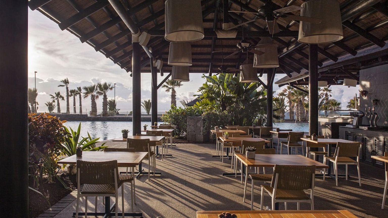 en restaurang med bord och stolar under ett halmtak med utsikt över en swimmingpool.