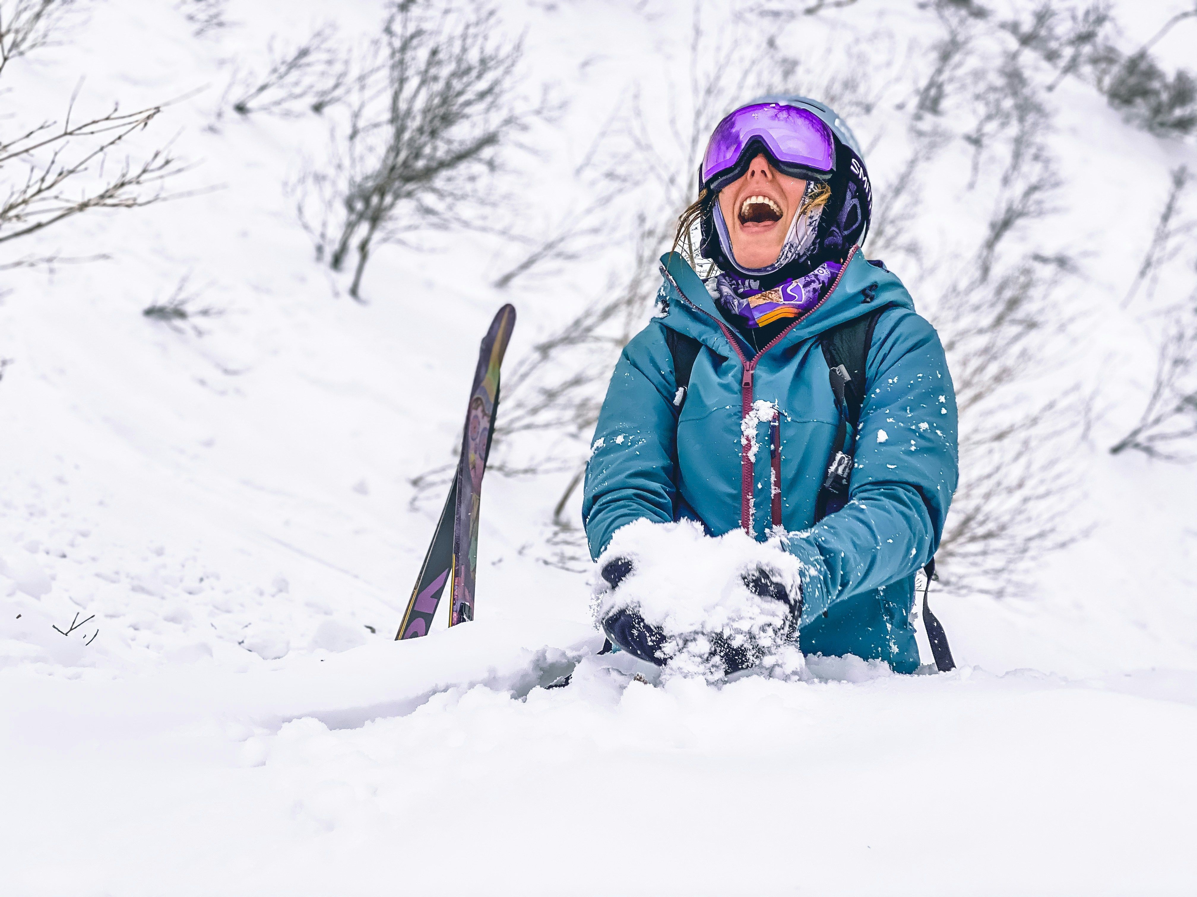 En glad skiløper i en turkis jakke og skibriller ler mens personen holder snø i et landskap med dyp snø.