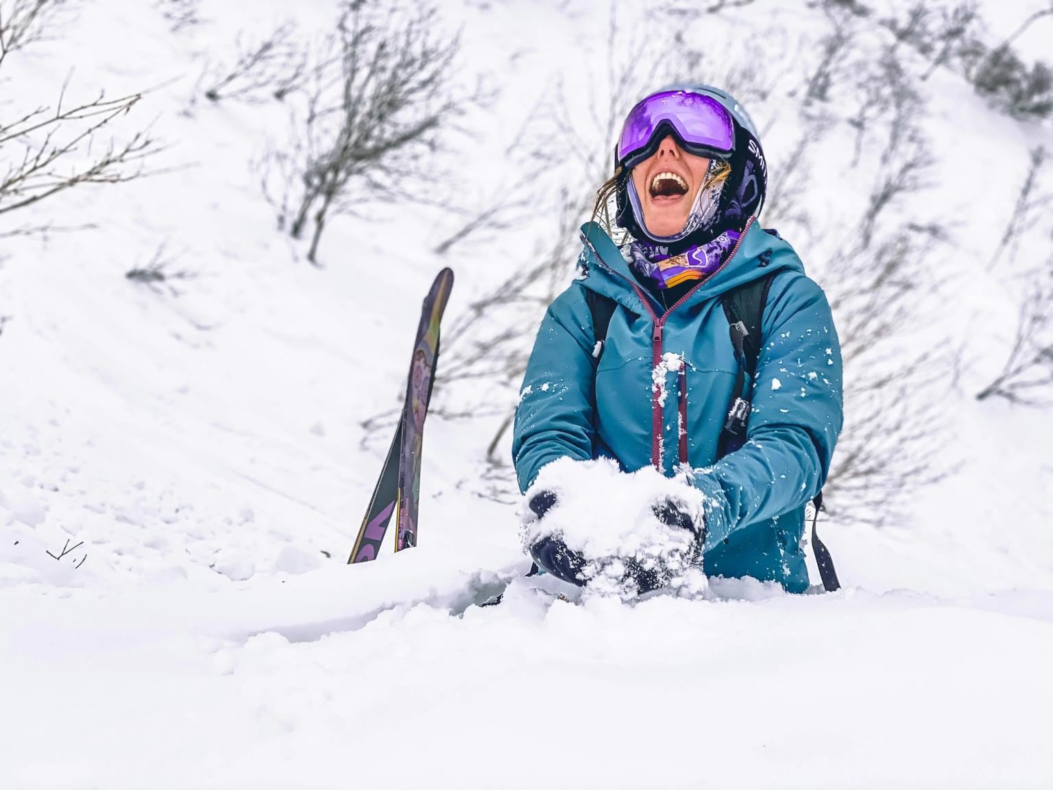 En glad skiløper i en turkis jakke og skibriller ler mens personen holder snø i et landskap med dyp snø.