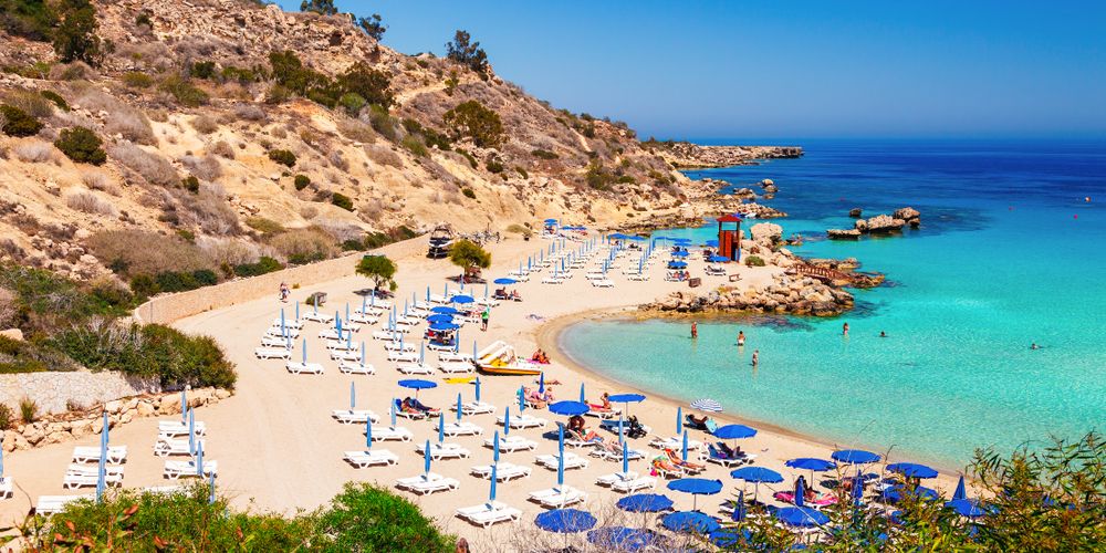 En pittoresk sandstrand med turkisfarget vann, omgitt av mange strandstoler og parasoller, med en steinete ås i bakgrunnen.