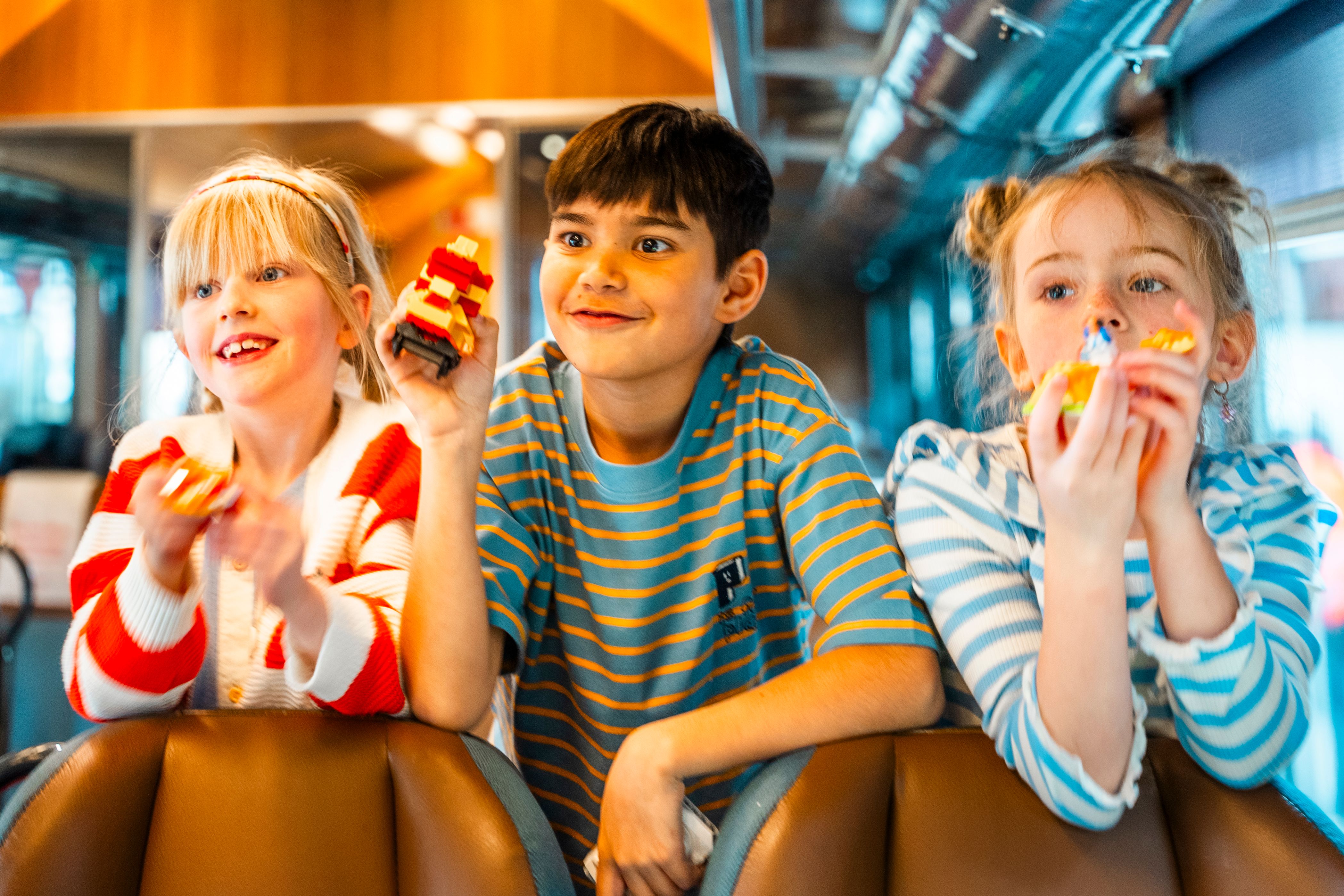 Tre glada barn håller små leksaker medan de tittar framåt på ett tåg.