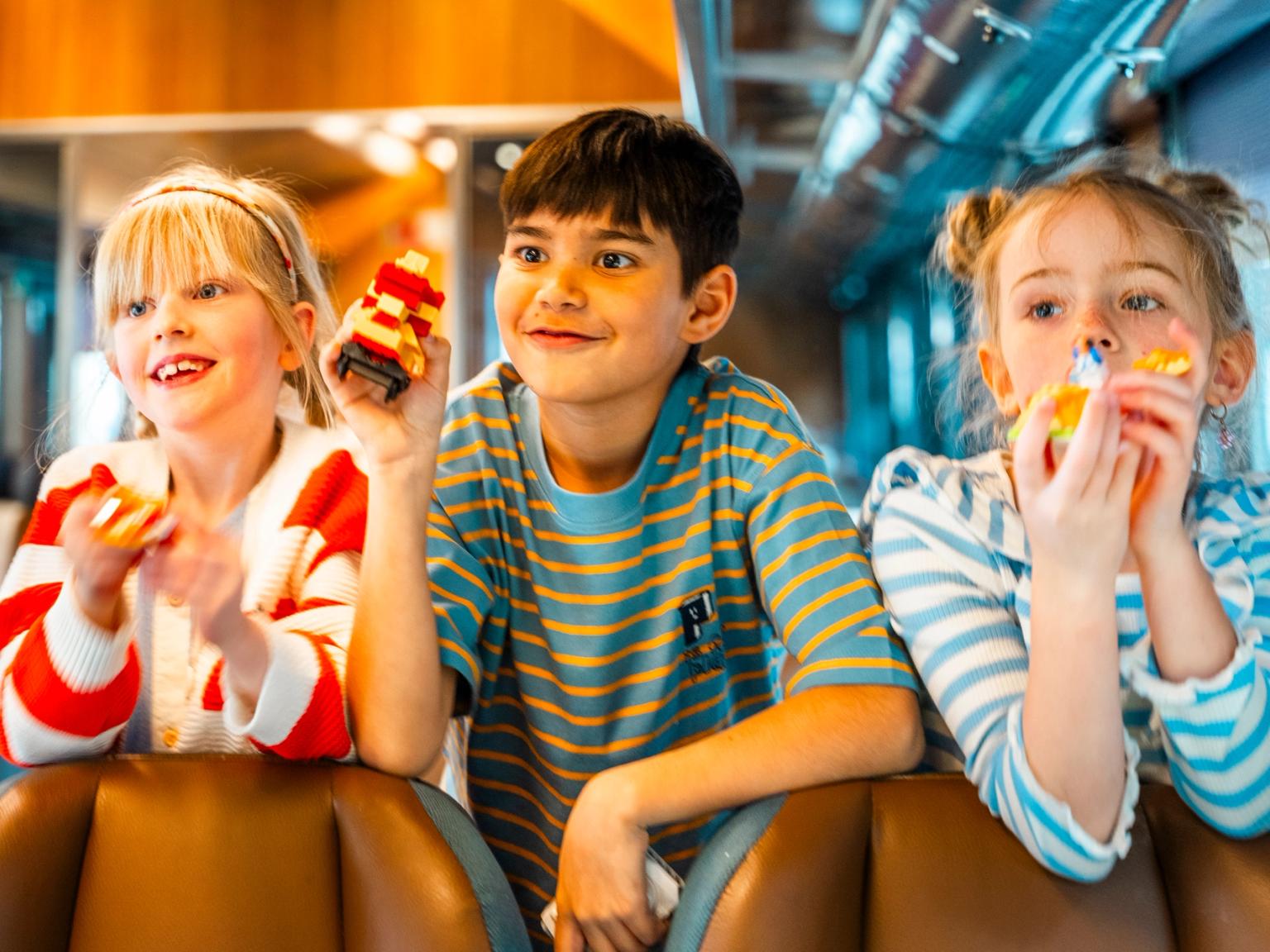 Tre glada barn håller små leksaker medan de tittar framåt på ett tåg.
