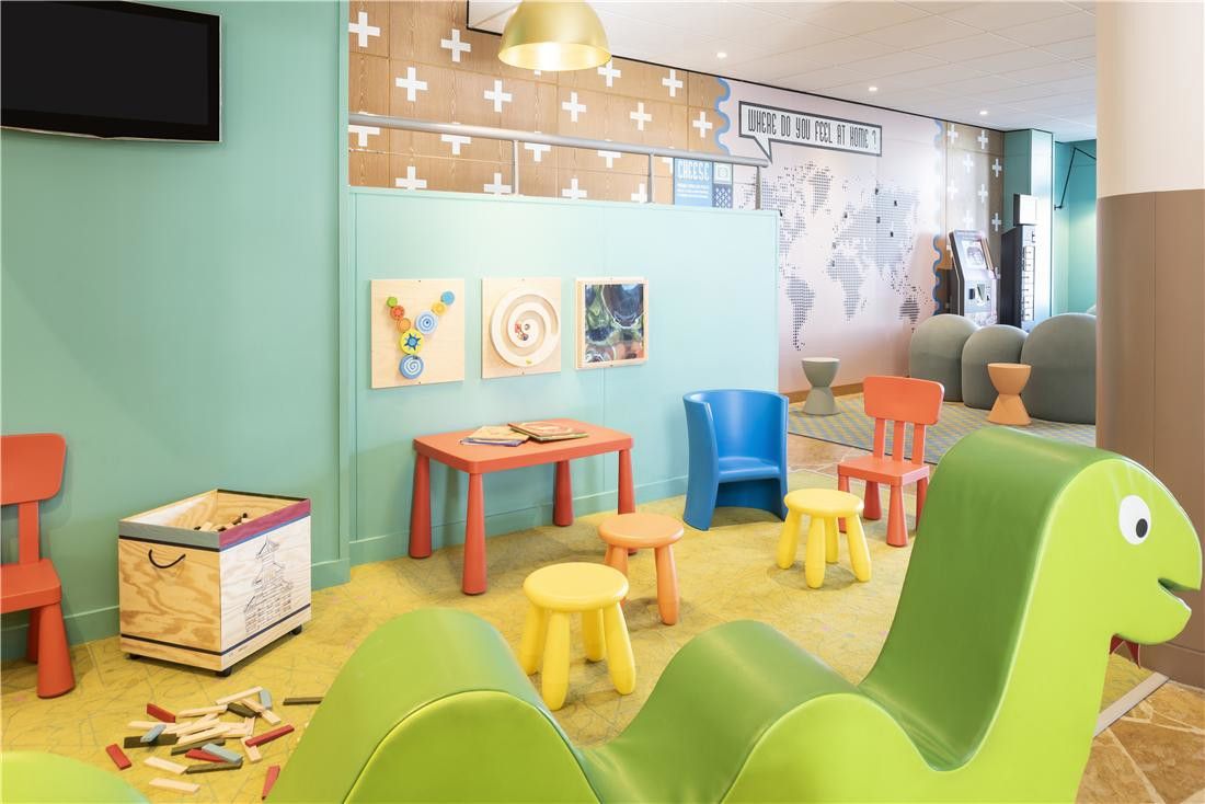 Et fargerikt lekeområde for barn med små bord og stoler, en grønn dinosaurformet myk lekestruktur og leker på gulvet. Adagio Aparthotel Val d'Europe nær Disneyland® Paris
