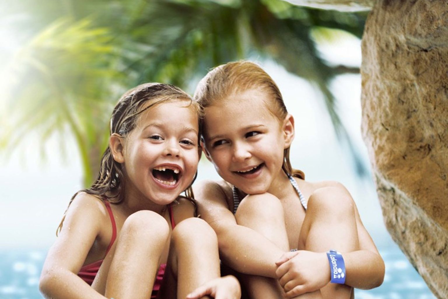 To glade unge piger med vådt hår og blå armbånd sidder på klipper i en tropisk grotte med vand og palmer bag dem. Lalandia Rødby