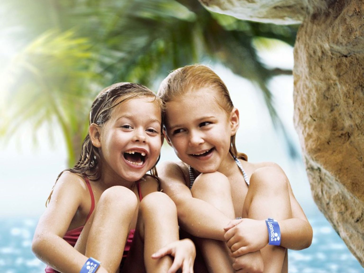 To glade unge jenter med vått hår og blå armbånd sitter på steiner i en tropisk grotte med vann og palmer bak dem. Lalandia Rødby
